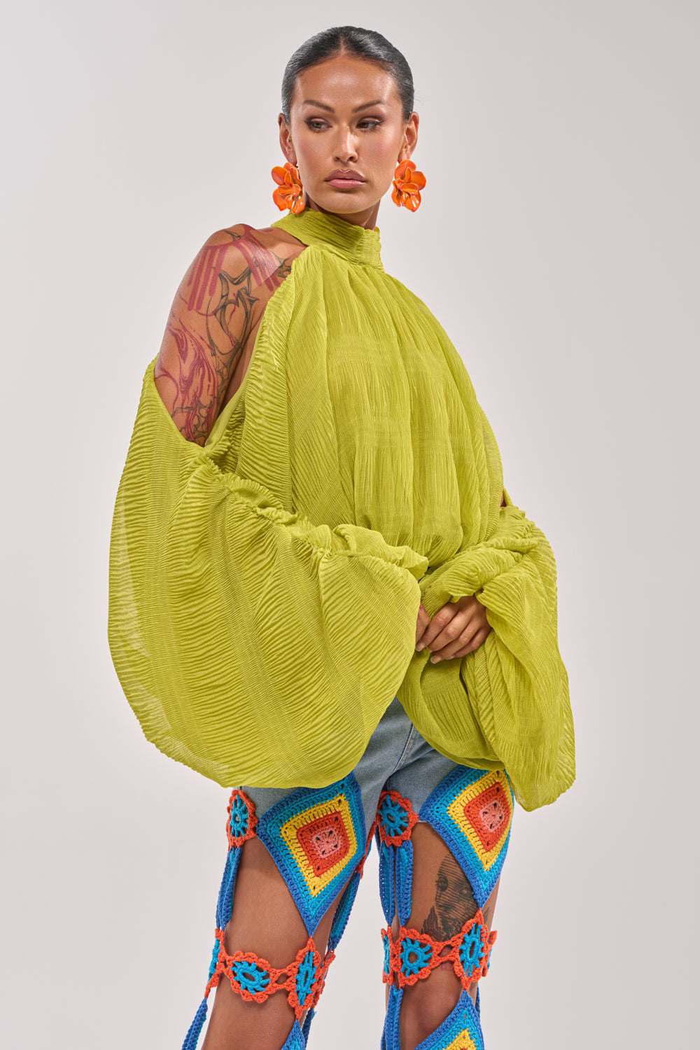 A person wears the NOT PLAYING BLOUSE in bright lime green with balloon sleeves, paired with large orange floral earrings and colorful crochet pants featuring blue, red, and yellow squares, posing against a plain background. 3