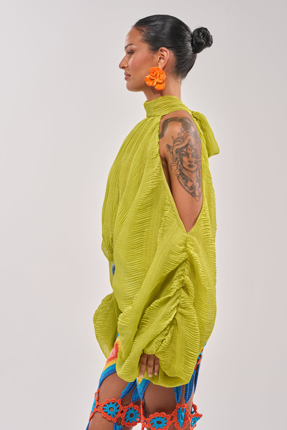 A woman with a black hair bun and orange flower earrings poses in profile, wearing the NOT PLAYING BLOUSE—a loose, textured green chiffon top with open sleeves—paired with a colorful crochet skirt. She displays a detailed upper arm tattoo. 4