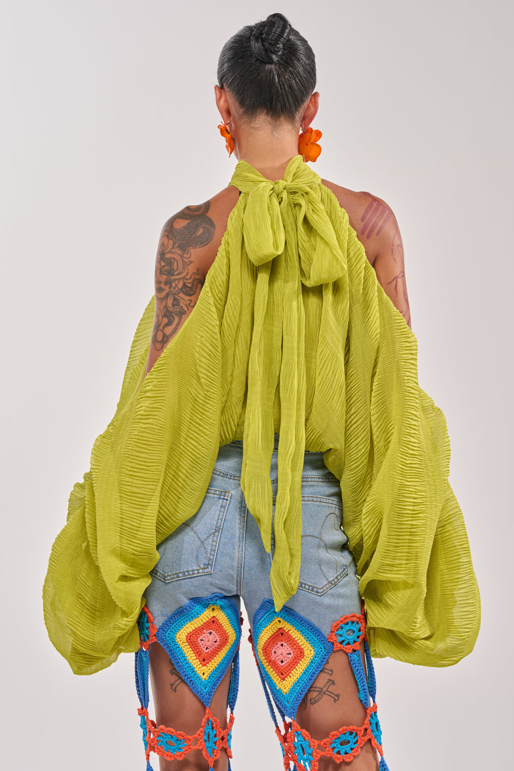 A person with short hair, showing tattoos, is seen from behind wearing the NOT PLAYING BLOUSE—a vibrant green chiffon top with billowy sleeves and a large bow—paired with denim shorts and colorful crochet leg accessories. 5