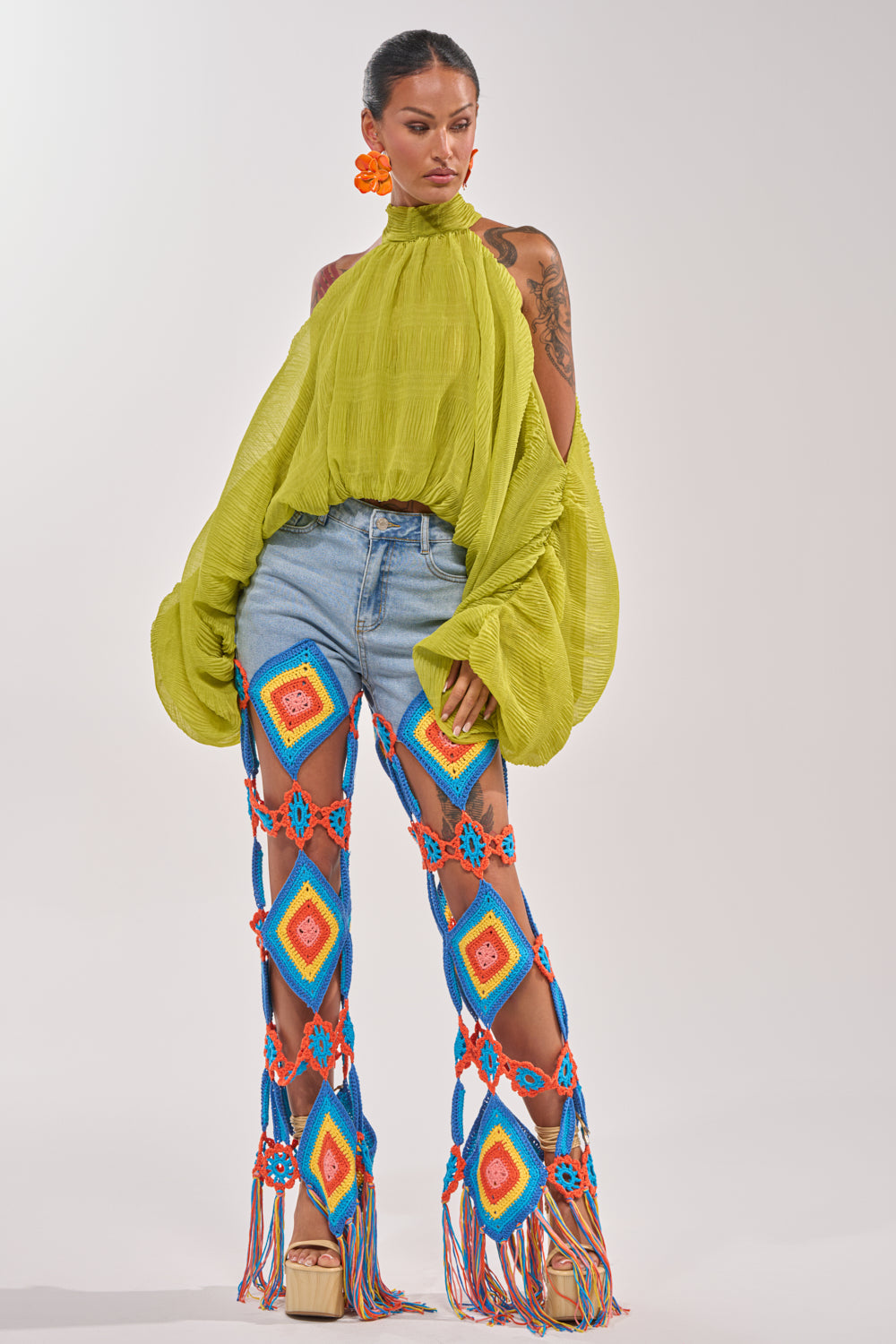 A woman models the NOT PLAYING BLOUSE in green chiffon with balloon sleeves, styled with light denim shorts, colorful crochet diamond-patterned fringe pants, beige platform sandals, and orange floral earrings. 6