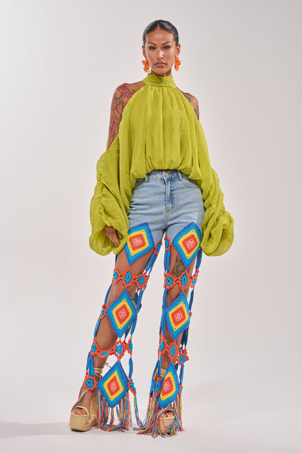 A woman models the NOT PLAYING BLOUSE, a bright green chiffon top with balloon sleeves, paired with denim shorts, colorful diamond-patterned blue crochet pants with fringe, platform sandals, and orange earrings for a vibrant look. 7