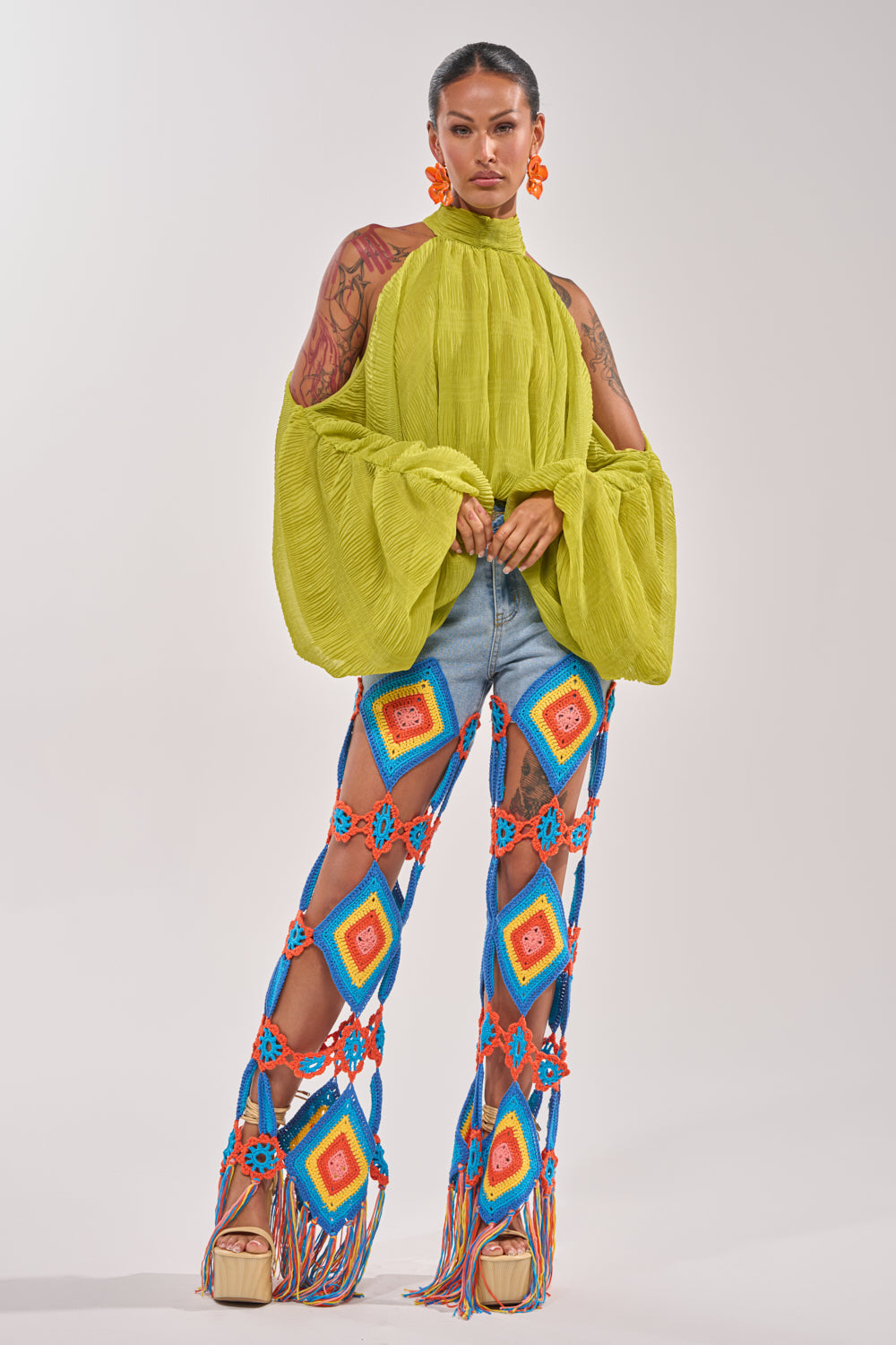 A woman in the NOT PLAYING BLOUSE, a green chiffon balloon sleeve top, pairs it with denim shorts and multicolored crochet lattice-style pants over platform sandals, standing against a plain backdrop. 8