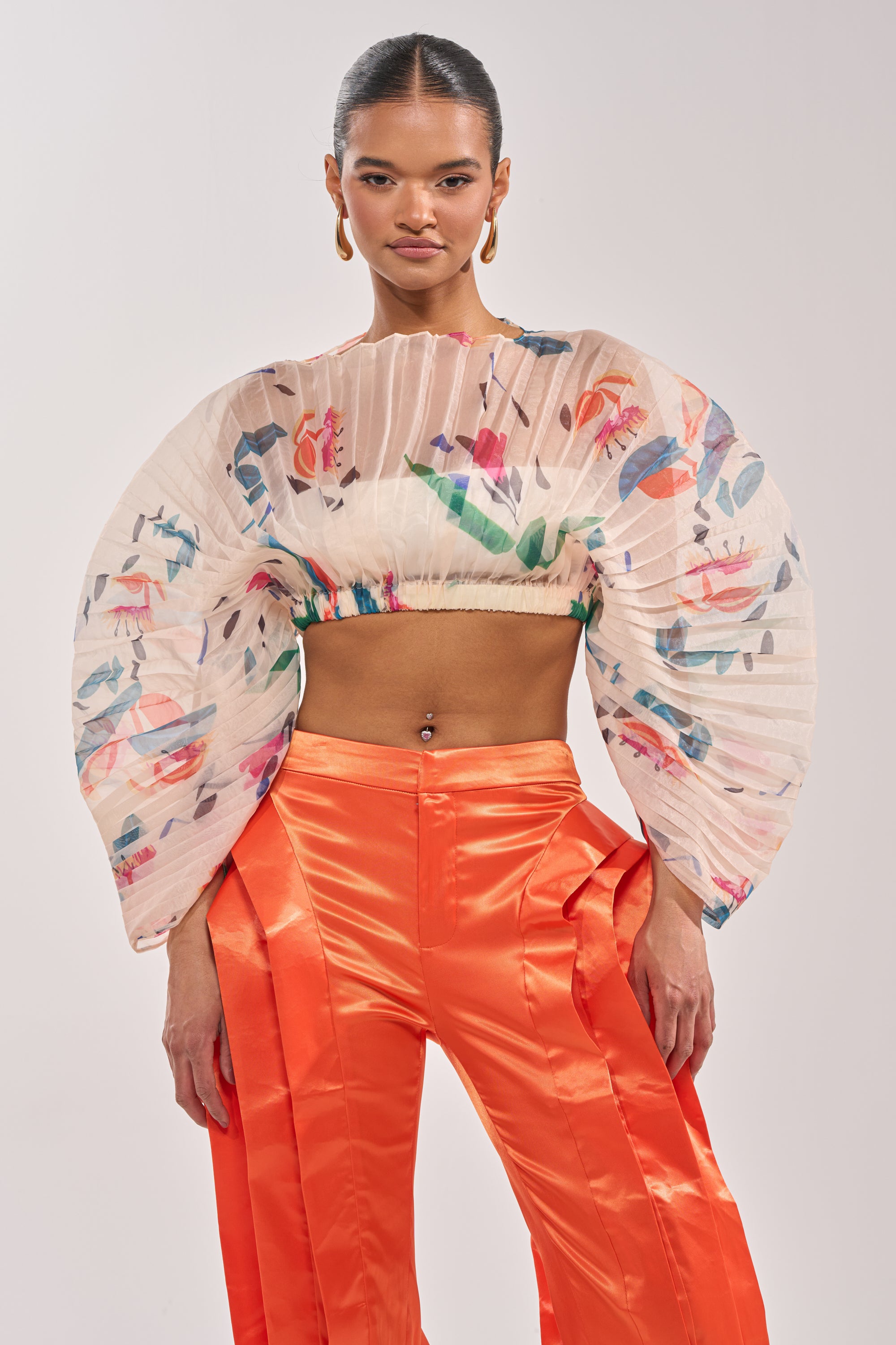 A woman stands confidently against a plain light background, wearing the CANDY RAIN PLEATED BLOUSE with colorful abstract patterns and sculptural sleeves, paired with bright orange pleated pants—a striking choice for chic occasions. 0