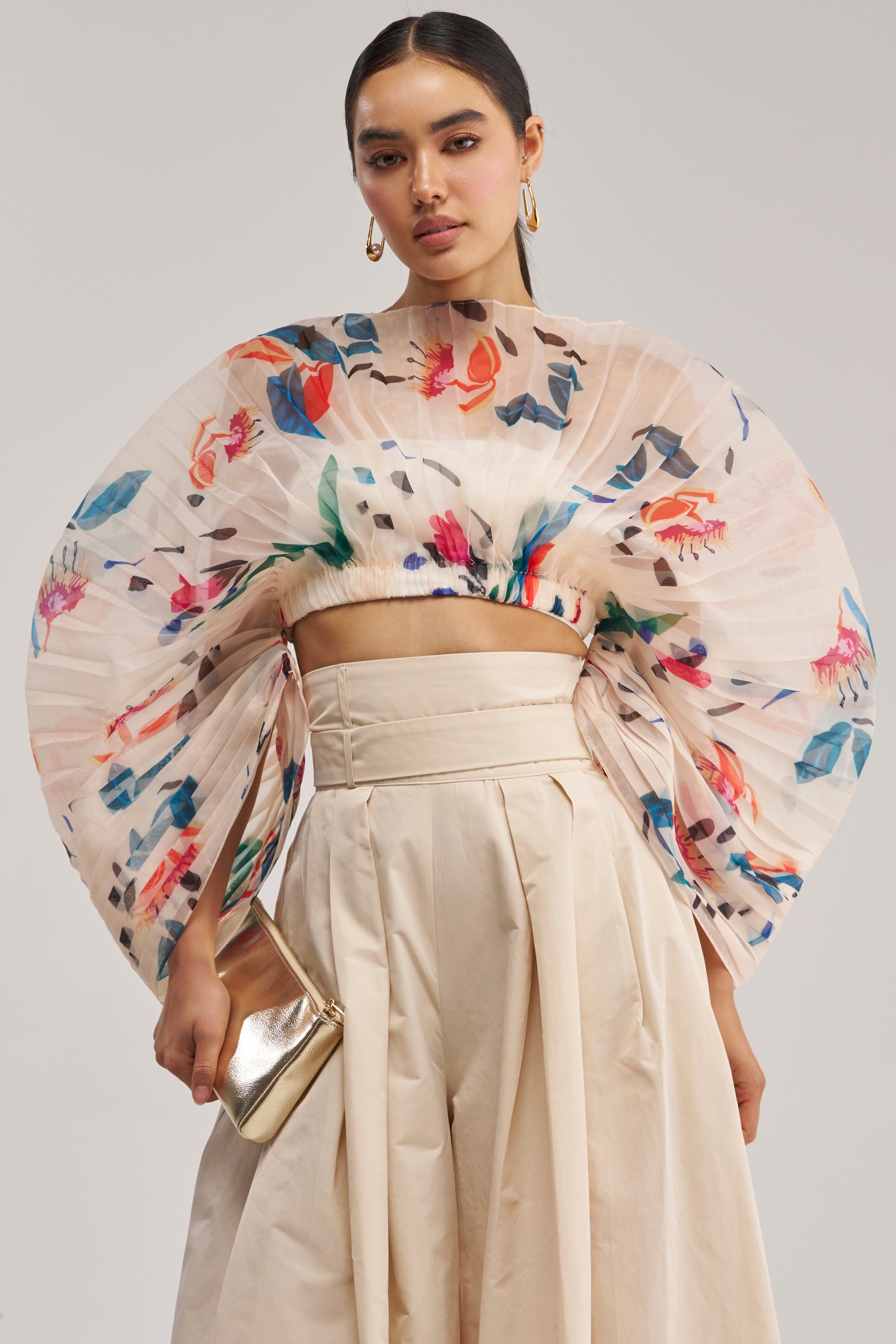 A woman wears the CANDY RAIN PLEATED BLOUSE—a spring-ready floral fan top with dramatic pleated sleeves—paired with a beige high-waisted skirt and accessorized with a gold clutch, slicked-back hair, and large hoop earrings. Perfect for occasion wear.