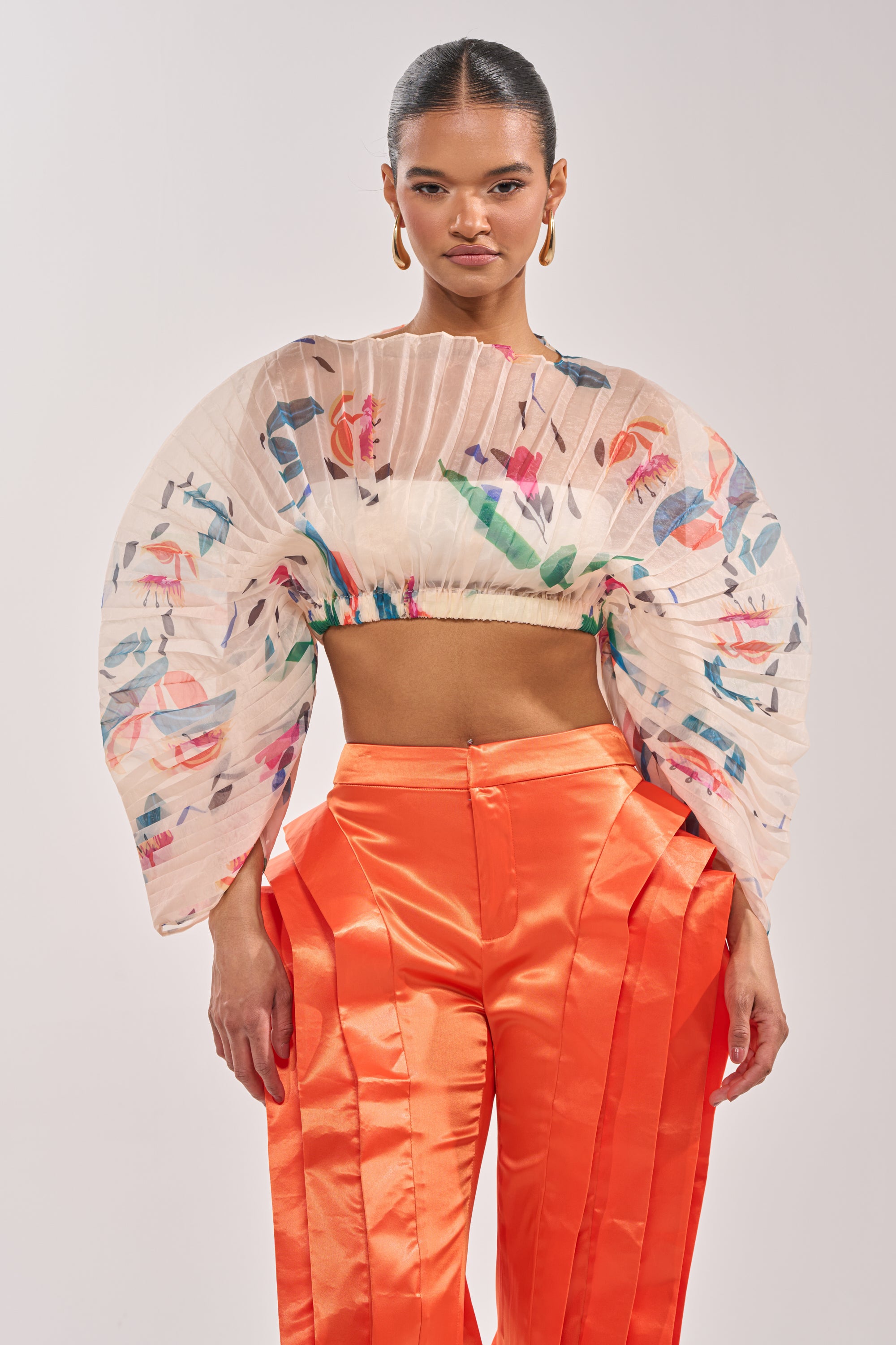 A woman showcases the CANDY RAIN PLEATED BLOUSE—a pleated floral, cropped top with exaggerated sleeves—paired with high-waisted, bright orange pleated pants for a bold occasion look against a neutral backdrop. 1
