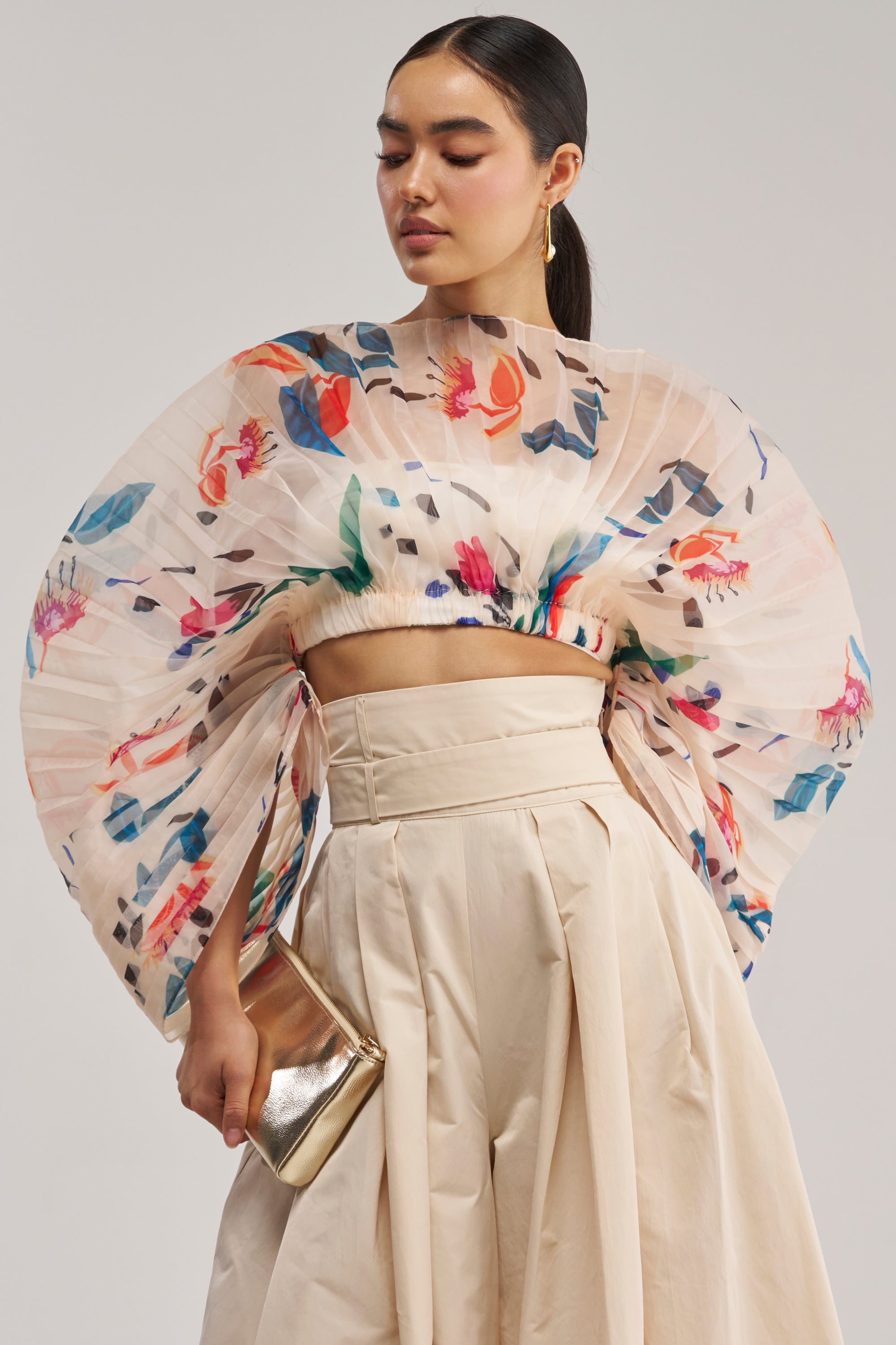 Wearing the CANDY RAIN PLEATED BLOUSE, this woman styles her occasion-ready look with voluminous sleeves, a high-waisted cream skirt, and a metallic clutch. She completes the ensemble with sleek hair and gold hoop earrings as she looks down gracefully.