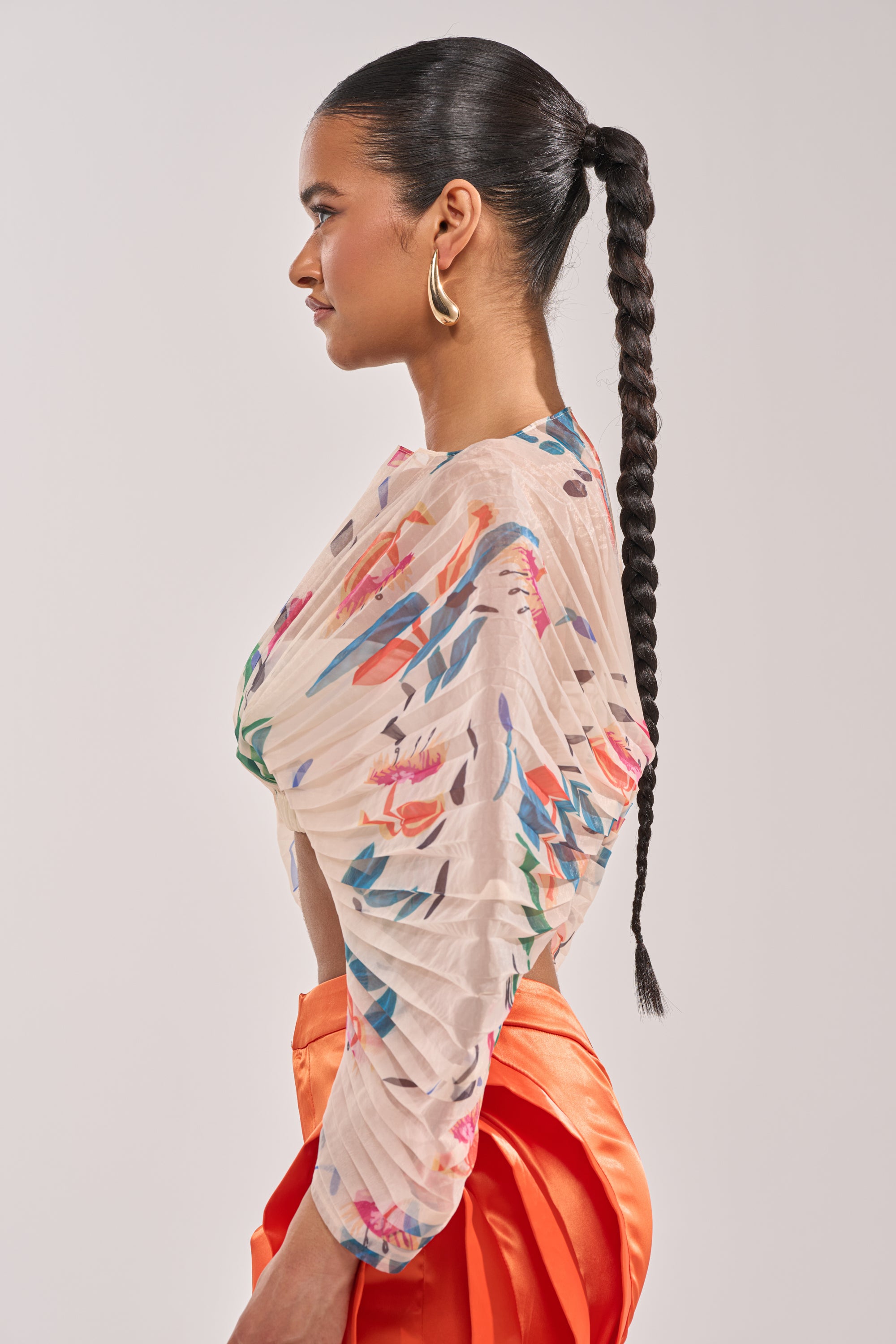A woman in profile, wearing large gold hoop earrings and the CANDY RAIN PLEATED BLOUSE with cropped sleeves, pairs it with bright orange high-waisted pants against a plain backdrop—ideal for stylish occasion wear. 2