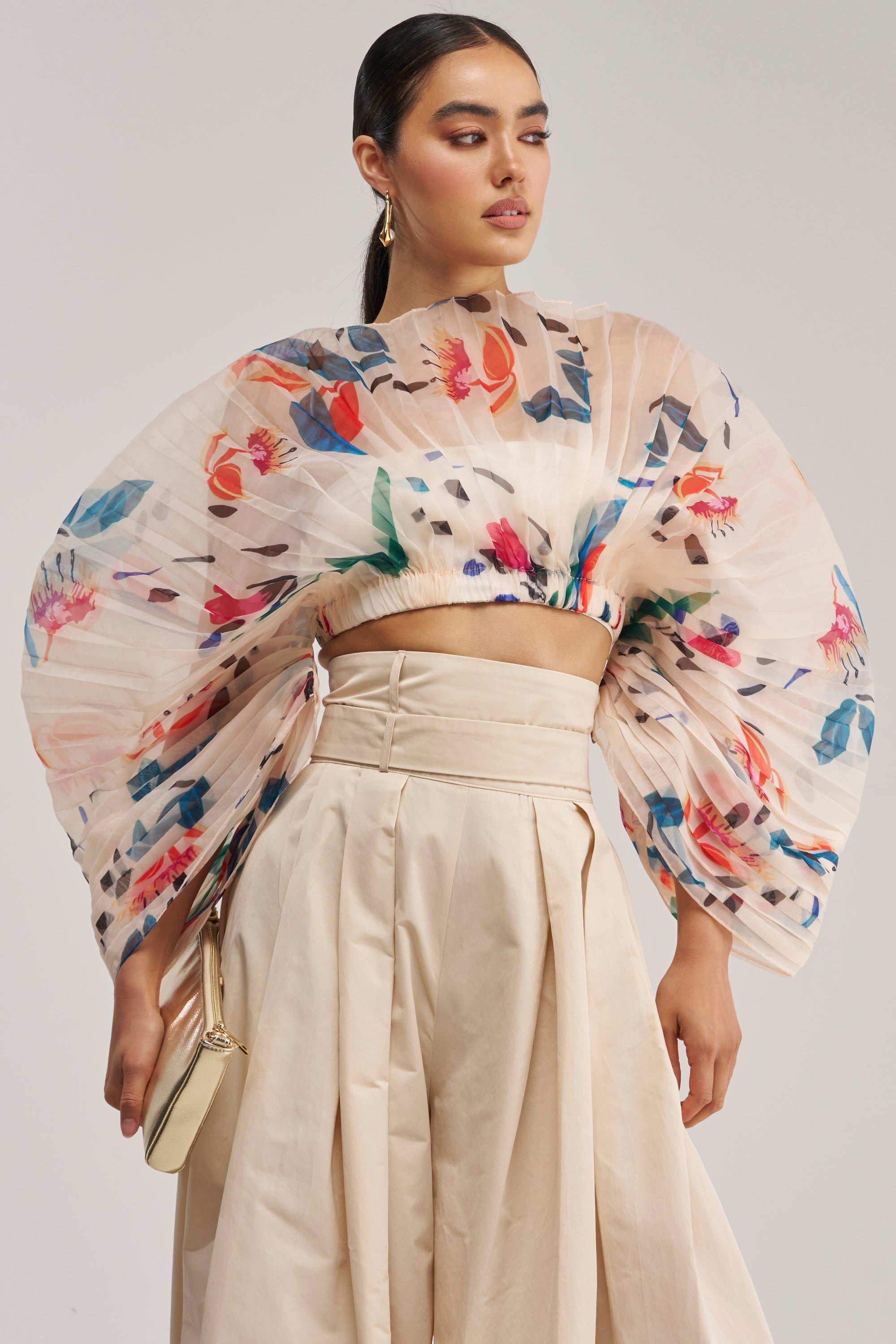 Wearing the CANDY RAIN PLEATED BLOUSE—a sheer, pleated floral fan top with voluminous sleeves—paired with high-waisted beige pants and a small beige purse, this woman gazes to the side against a plain background for an effortlessly spring-ready look that’s perfect for any occasion.