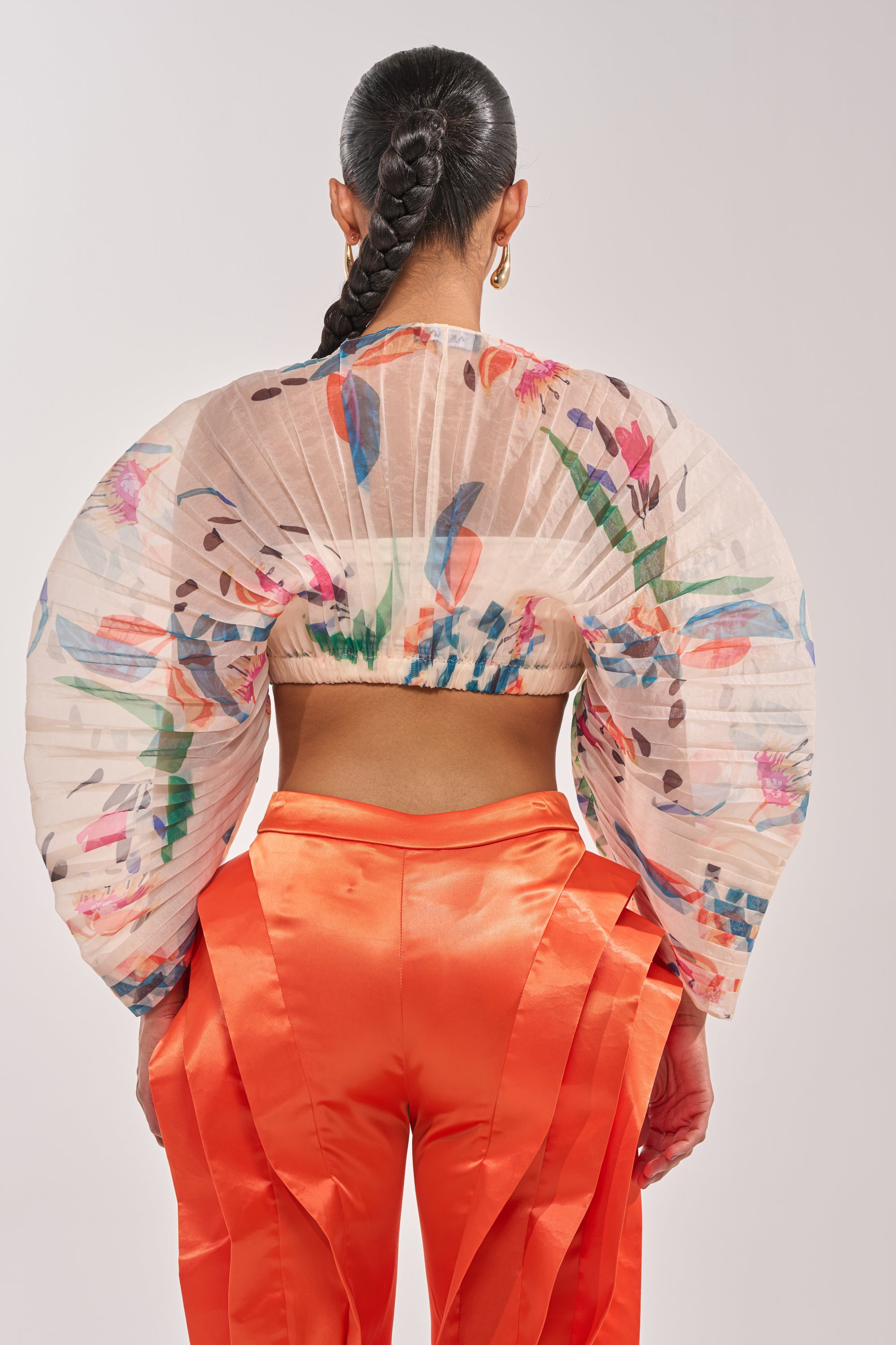 A person with a long braided ponytail faces away, wearing the CANDY RAIN PLEATED BLOUSE—sheer, cropped, with rounded sleeves—and vibrant orange satin pants for an eye-catching occasion look. 3