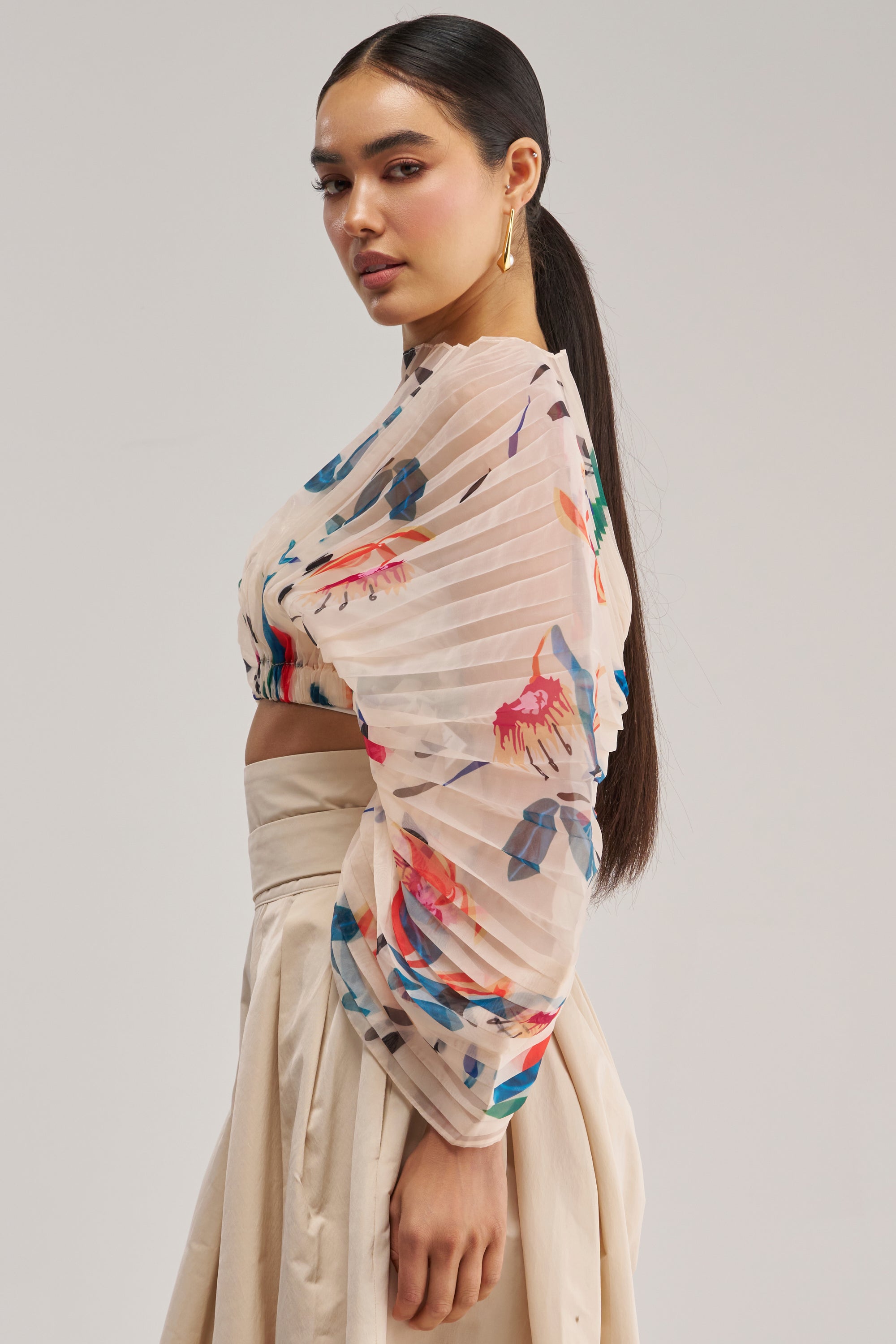 A woman in profile wears the CANDY RAIN PLEATED BLOUSE, featuring spring-ready floral pleats and voluminous sheer sleeves, paired with a beige high-waisted skirt. Her long dark hair is styled in a low ponytail, and she accessorizes with gold earrings against a plain light background.