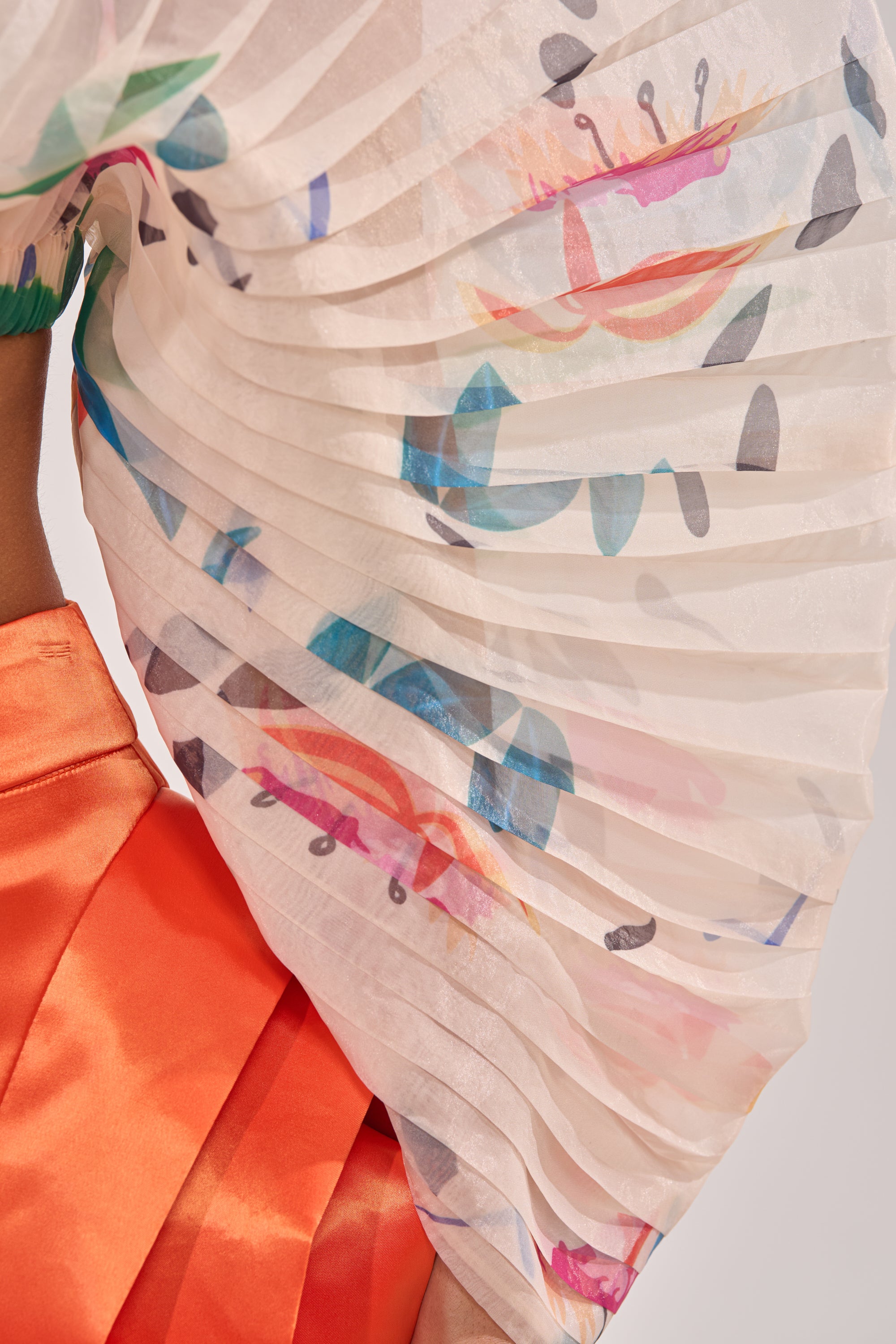 A close-up of a person wearing the CANDY RAIN PLEATED BLOUSE—a sheer, pleated floral top with vibrant abstract patterns—styled elegantly for occasion wear and draped over the shoulder. 7