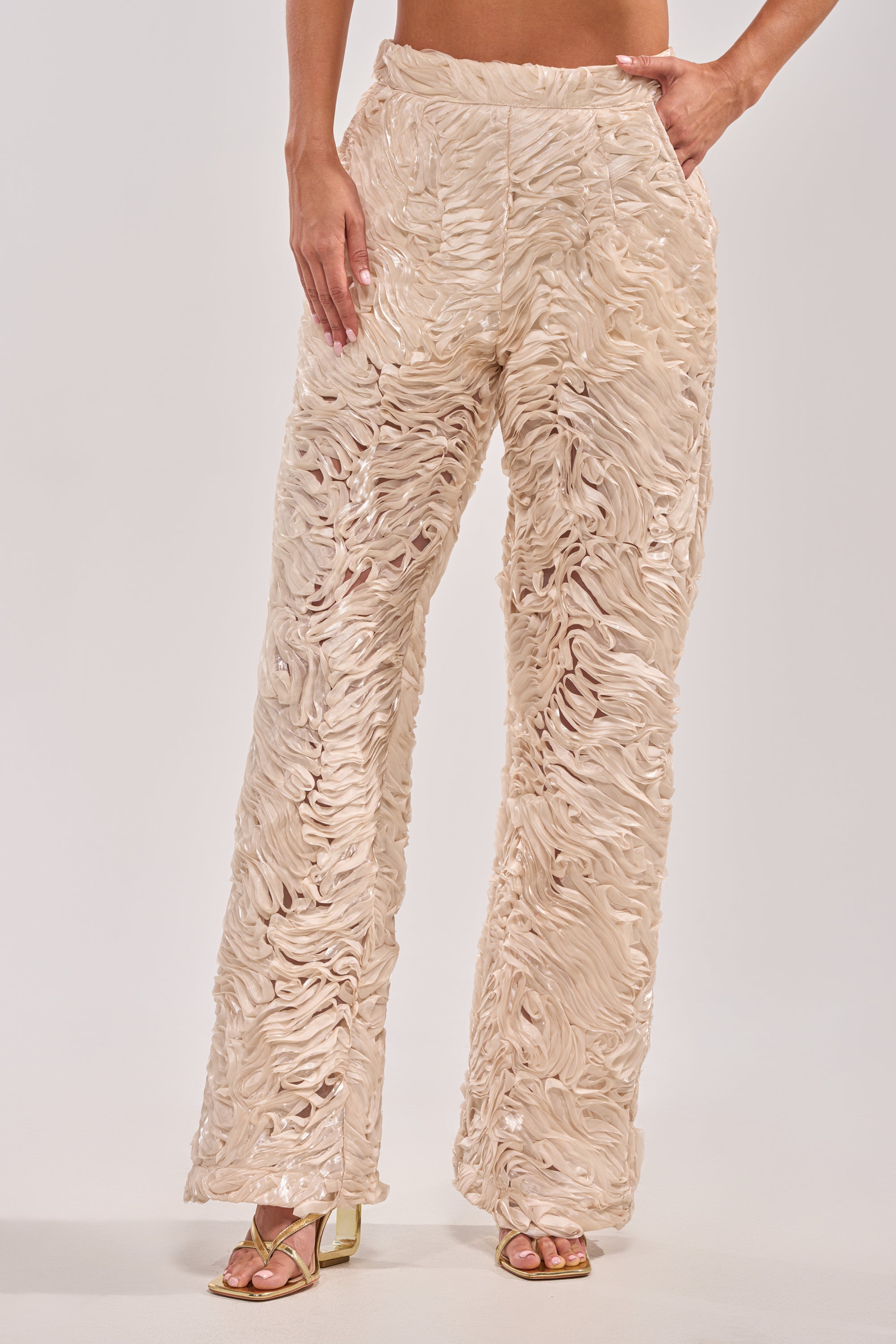 A person in the WAVY AND COOL TROUSER—high-waisted, wide-leg ivory trousers with a shiny, wavy textured pattern—paired with gold open-toe heels poses against a plain white background, ideal for chic white party looks.