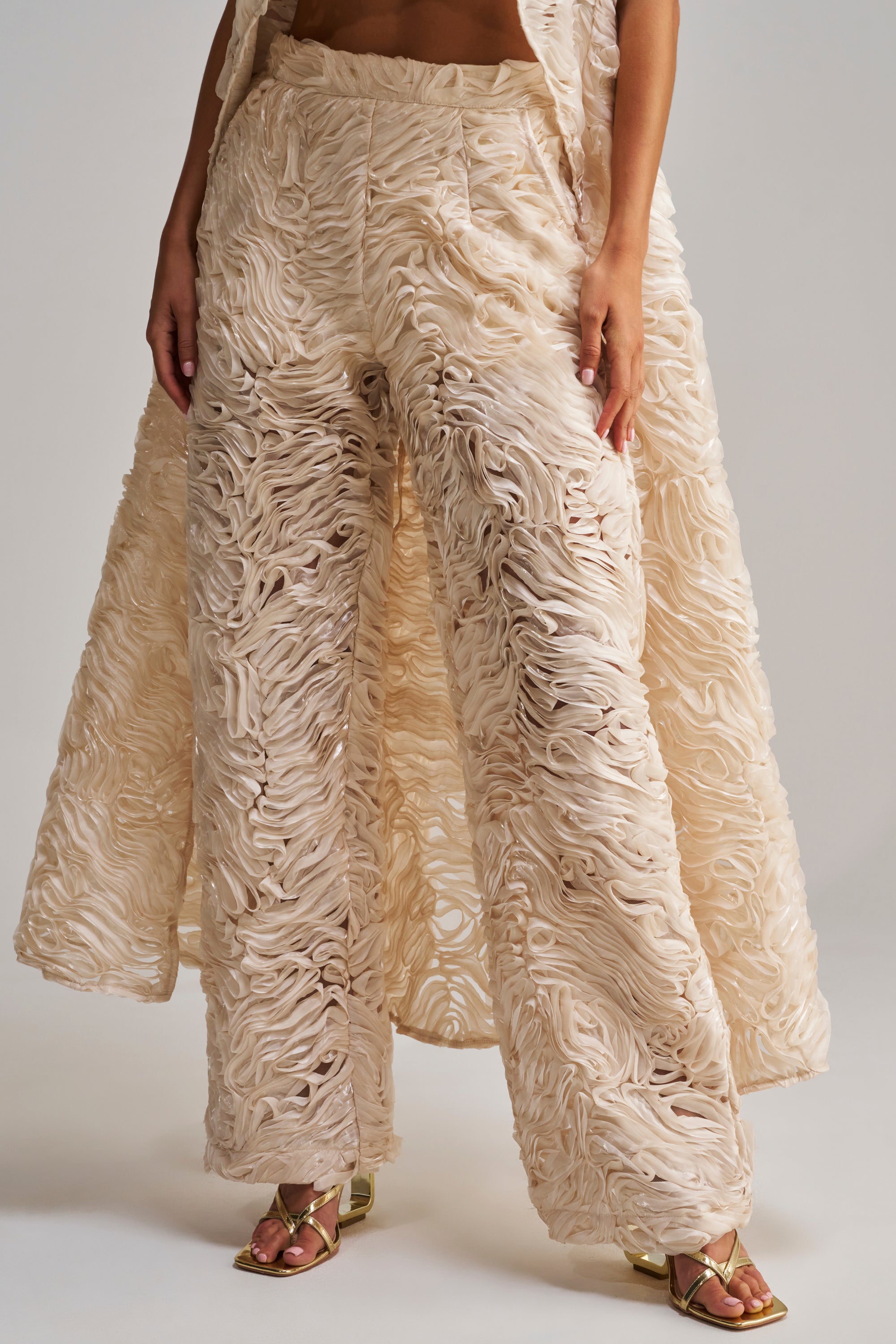A person wears the WAVY AND COOL TROUSER, featuring a wide-leg beige textured design with layered ruffles and metallic heeled sandals. The matching long coat drapes behind, hands at their sides. The image is cropped at the torso.
