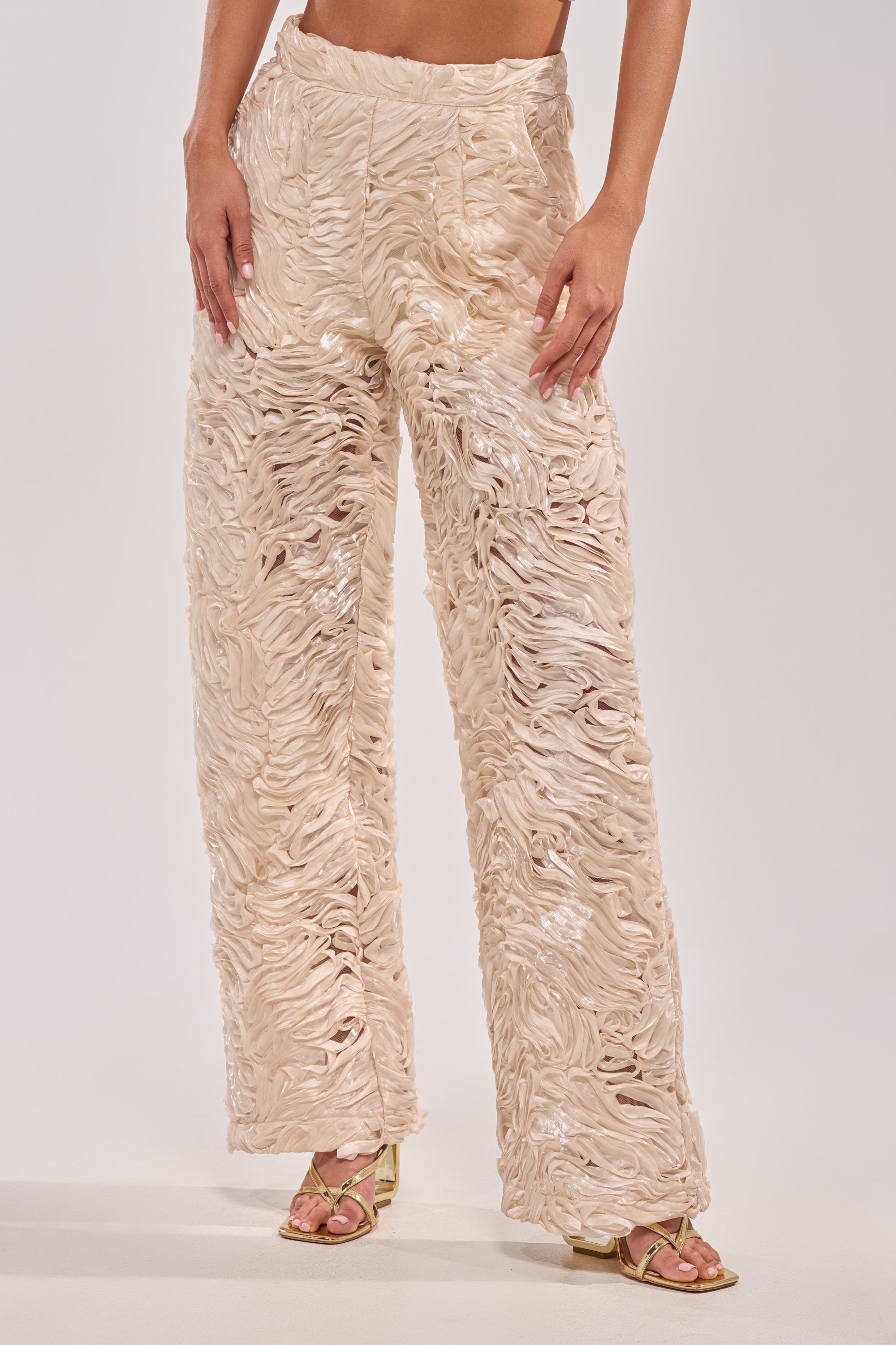 Wearing WAVY AND COOL TROUSER featuring an ivory hue with wavy, textured details and shiny accents, paired with light open-toe heeled sandals—an elegant choice for white party outfits. Only the lower torso and legs are shown.