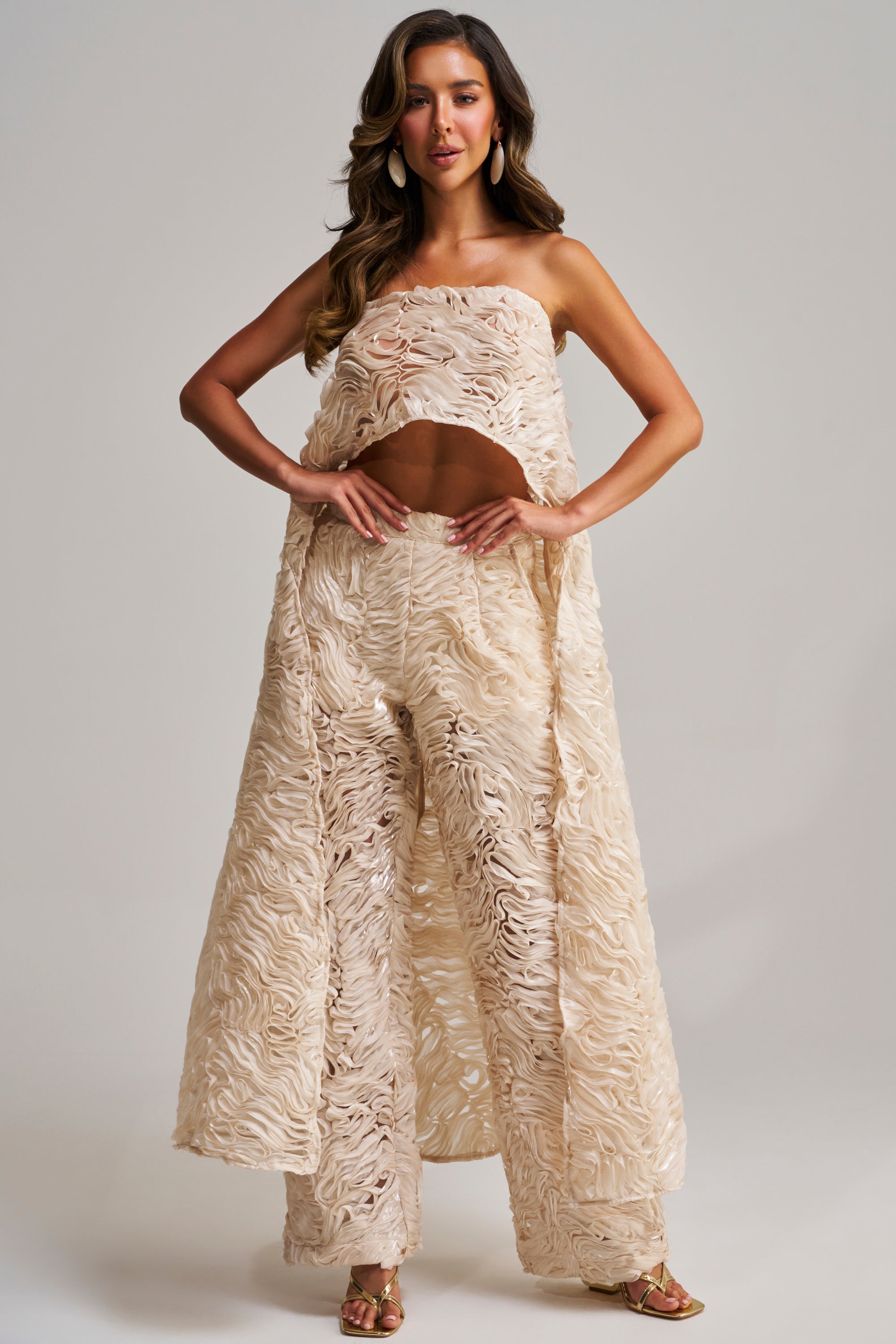 A woman poses in the MAEVE SLEEVELESS BLOUSE, a cream-colored, textured two-piece with wide-leg pants and a flowing overlay—an elegant white party outfit. She pairs it with gold high-heeled sandals and loose, wavy hair.