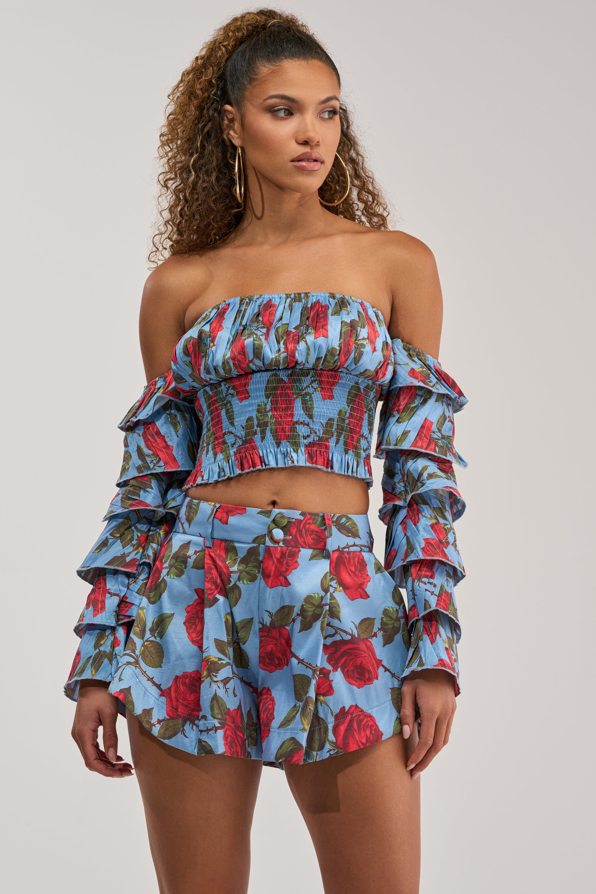 A woman wearing the ROSES FOR EVERYONE OFF THE SHOULDER BLOUSE with a smocked bodice and cropped hemline, paired with high-waisted shorts featuring a red rose print, stands against a plain background.