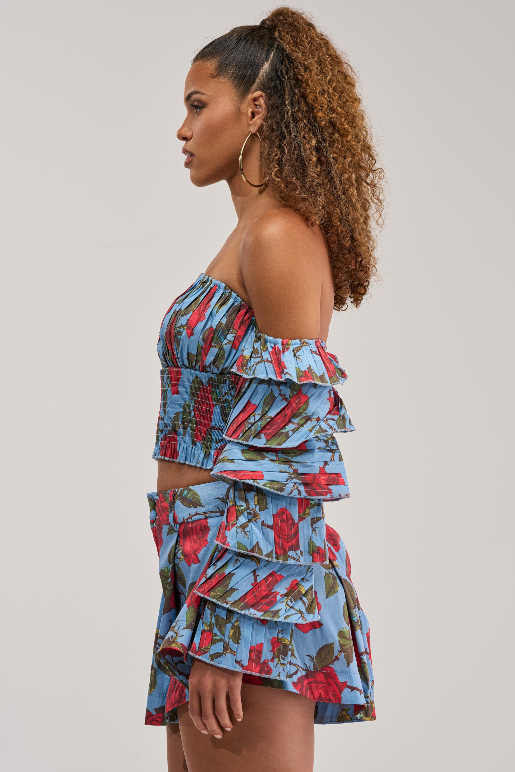 A woman with curly hair styled in a high ponytail is pictured in profile against a plain background, wearing the ROSES FOR EVERYONE OFF THE SHOULDER BLOUSE—a blue floral top with a smocked bodice—paired with a matching skirt featuring ruffled layers and a cropped hemline.