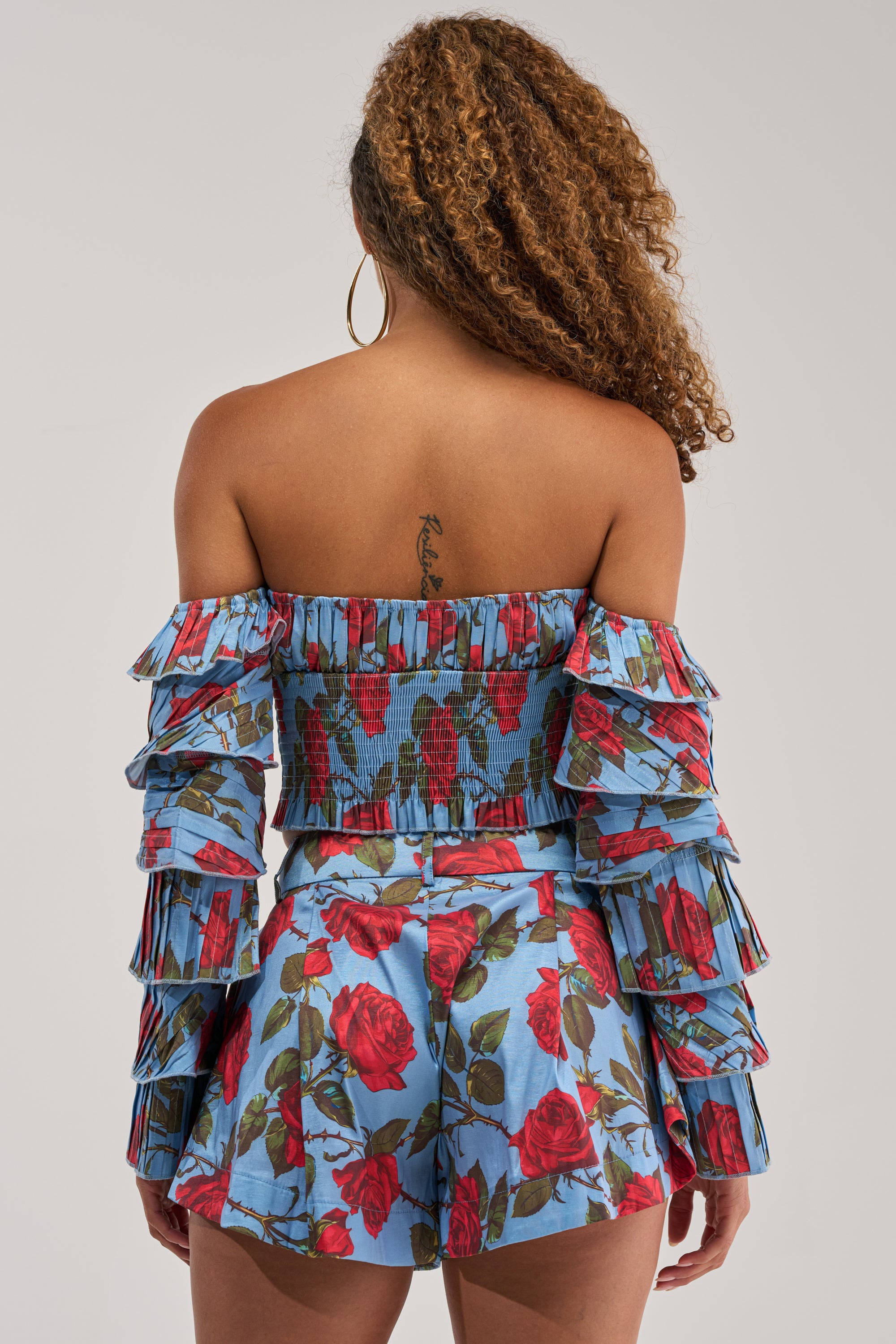 A woman with curly hair faces away, showcasing the ROSES FOR EVERYONE OFF THE SHOULDER BLOUSE—a blue top featuring a red floral print and pleated sleeves. She has a tattoo on her upper back and wears large hoop earrings.
