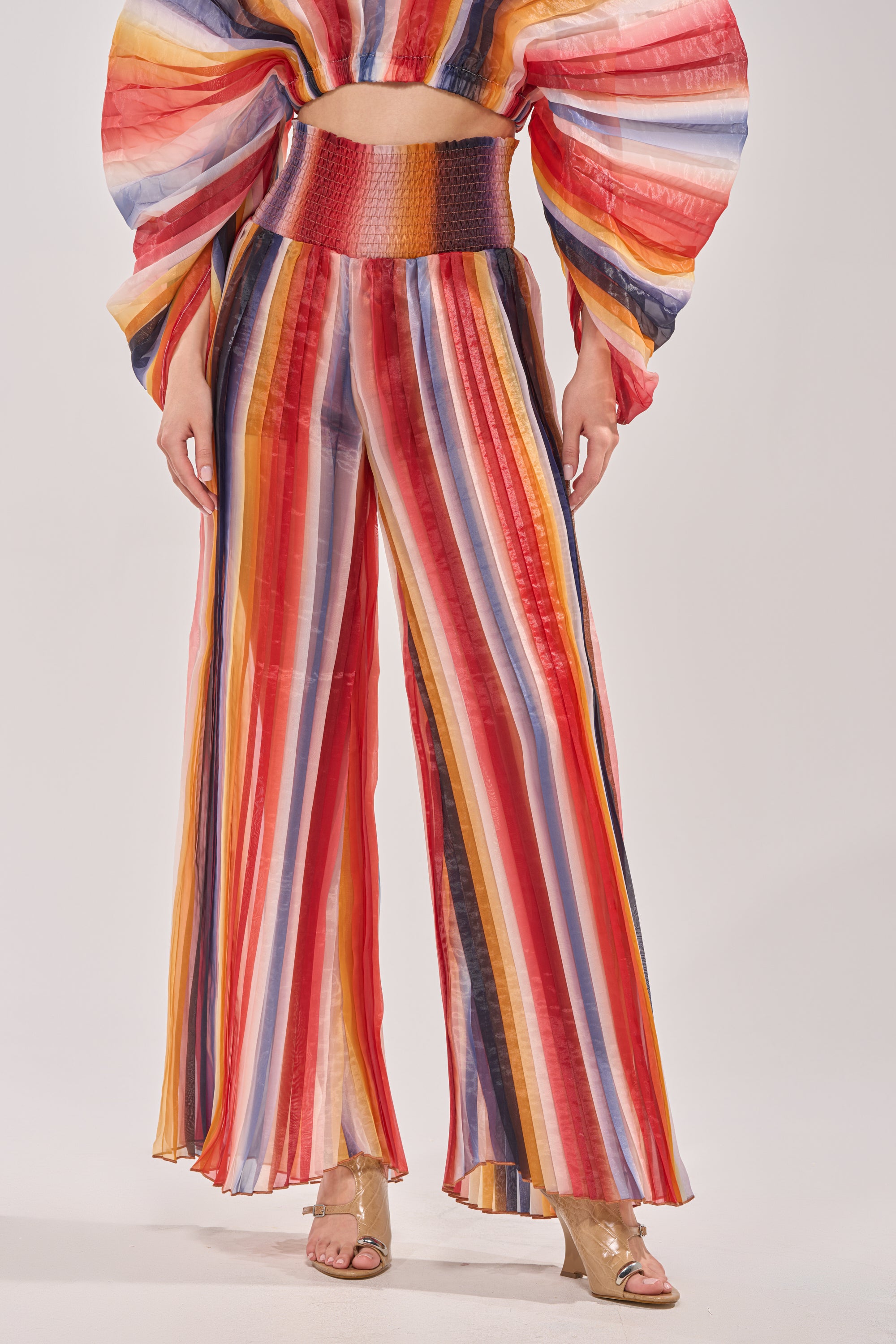 A person wears the FANNED OUT CHIFFON PANT—multicolor, wide-leg, high-rise chiffon pants—paired with gold open-toe heels and a matching puff-sleeve cropped top that shows a hint of midriff.
