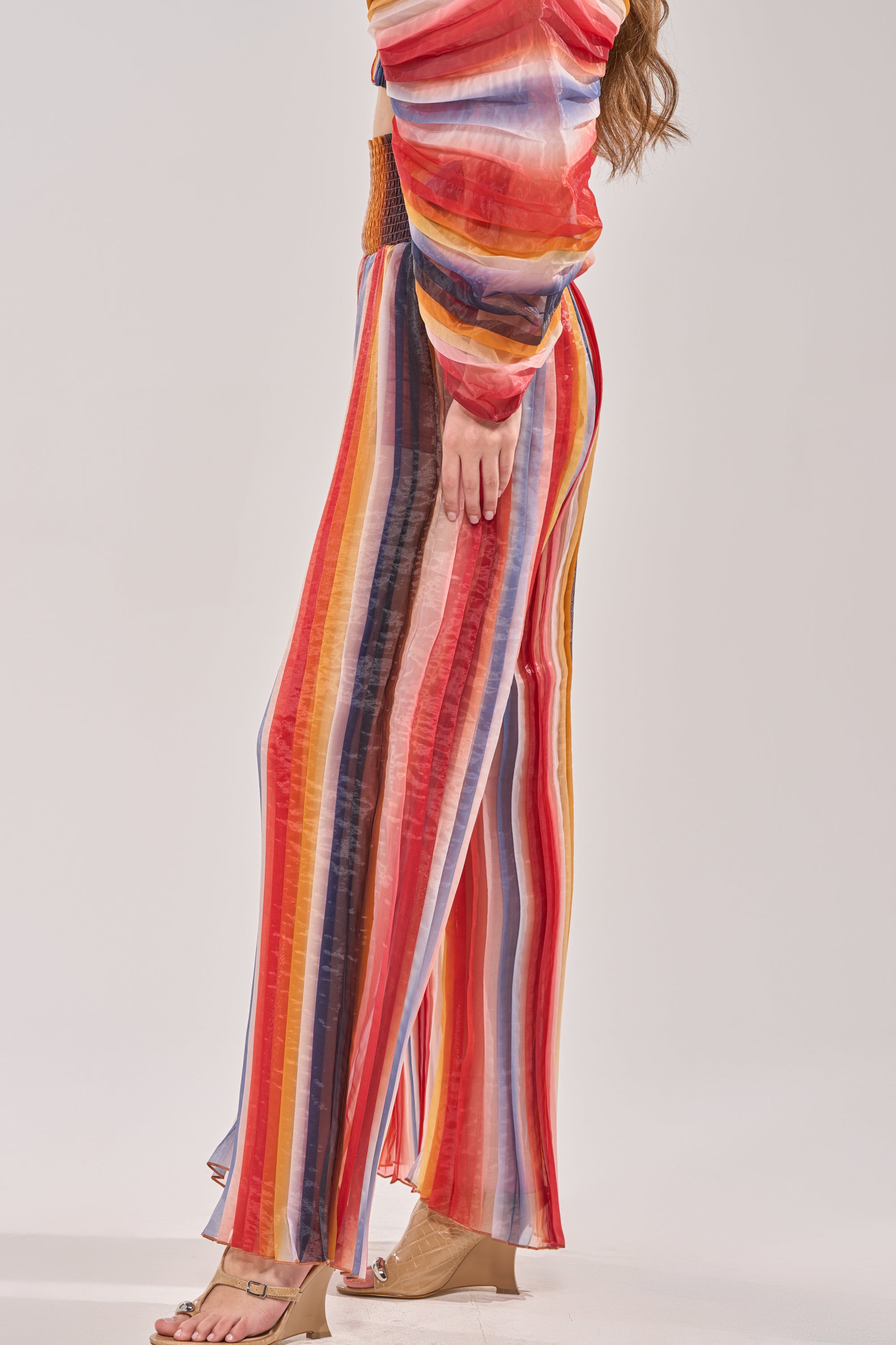 A person wears the FANNED OUT CHIFFON PANT—high-rise, wide-leg chiffon pants with vertical rainbow stripes—and a matching long-sleeve top, standing barefoot on a light background.