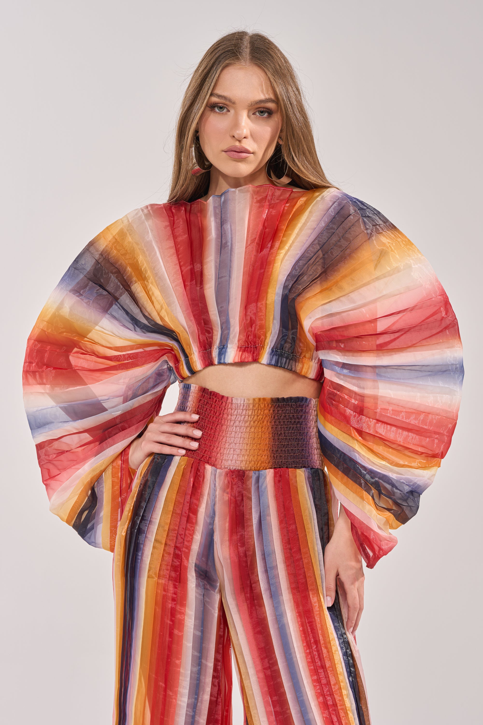 A woman models the FANNED OUT CHIFFON BLOUSE, a colorful sheer top with wide fan sleeves, paired with high-waisted pants featuring bold red, yellow, orange, blue, and white stripes. 0