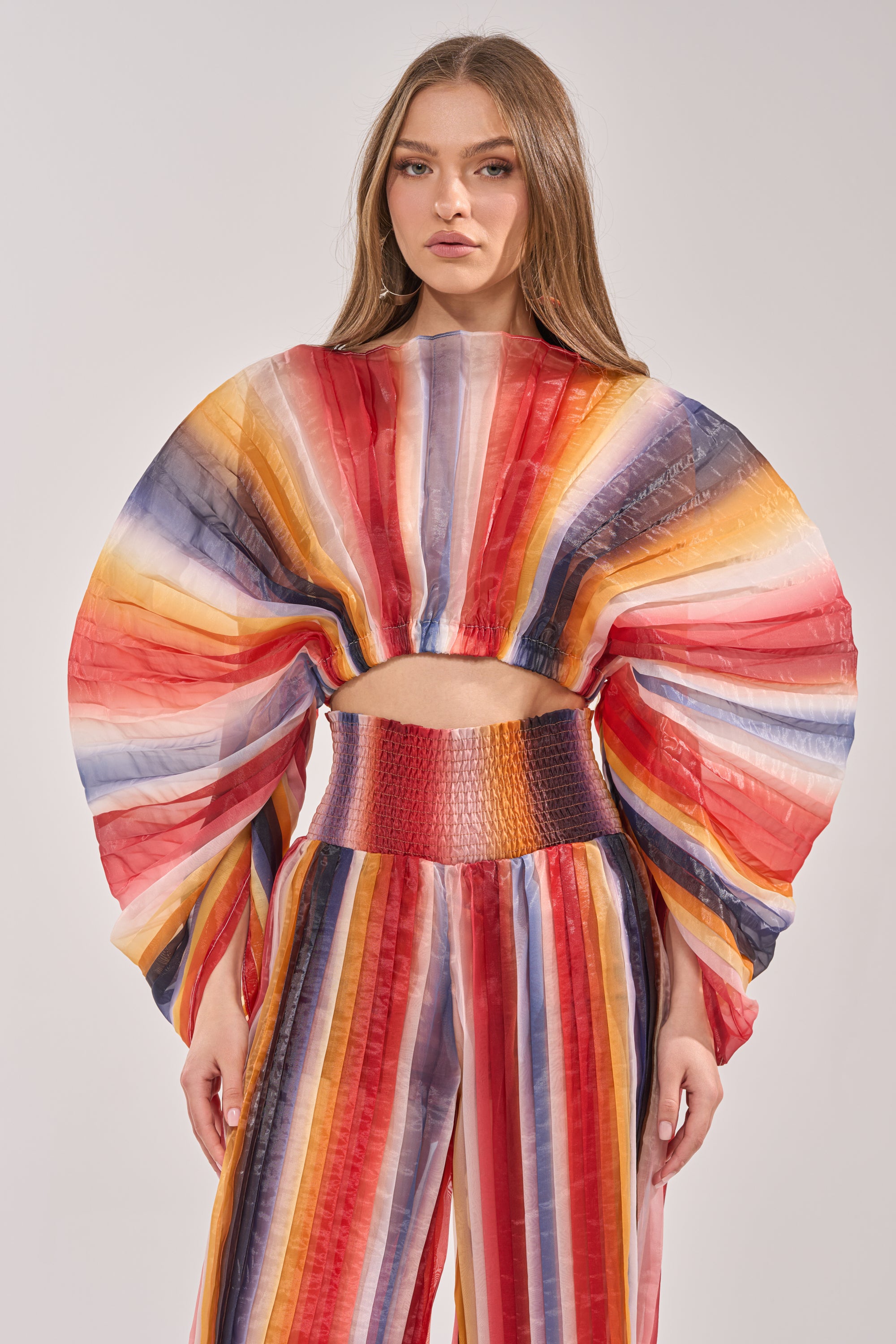 A woman wears the FANNED OUT CHIFFON BLOUSE— a multicolor, cropped blouse with voluminous semi-sheer sleeves and vertical stripes in red, orange, yellow, blue, and white — paired with matching high-waisted pants. 1