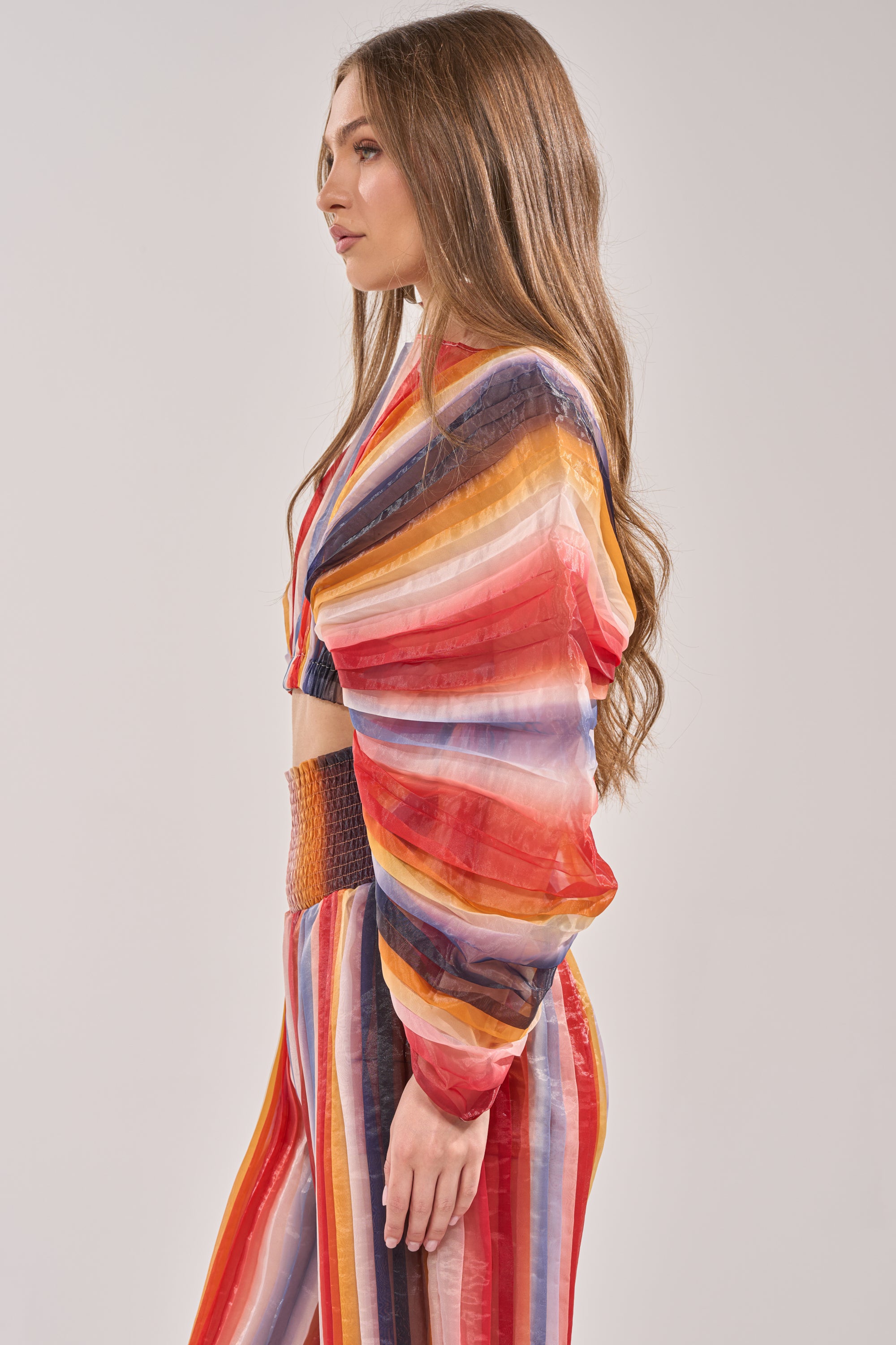 A woman with long brown hair models the FANNED OUT CHIFFON BLOUSE—featuring oversized fan-shaped sleeves—in profile with high-waisted pants against a plain light background, creating a bold summer statement look. 2