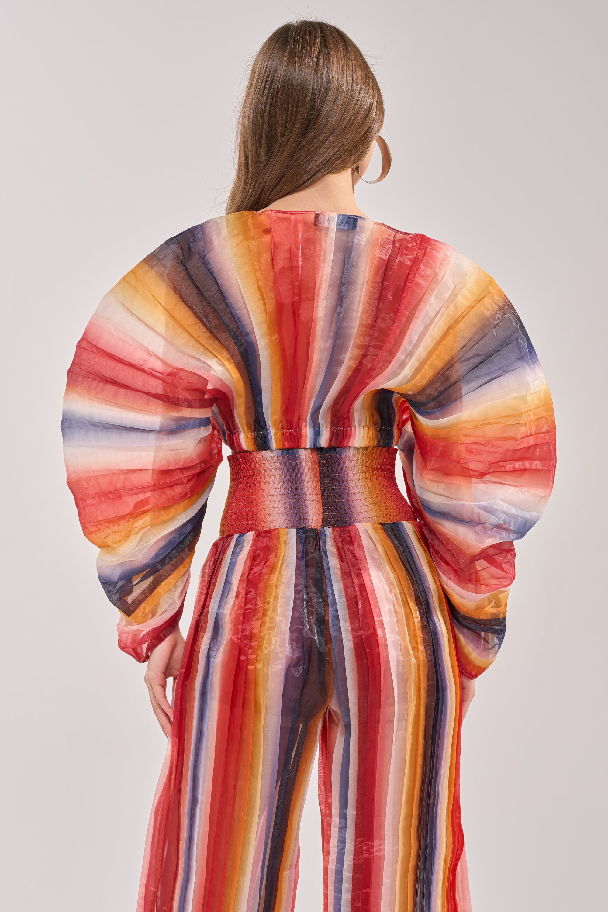 A person with long hair is shown from the back in the FANNED OUT CHIFFON BLOUSE, a colorful top featuring wide, dramatic sleeves and vibrant vertical rainbow stripes, set against a plain light background. 3