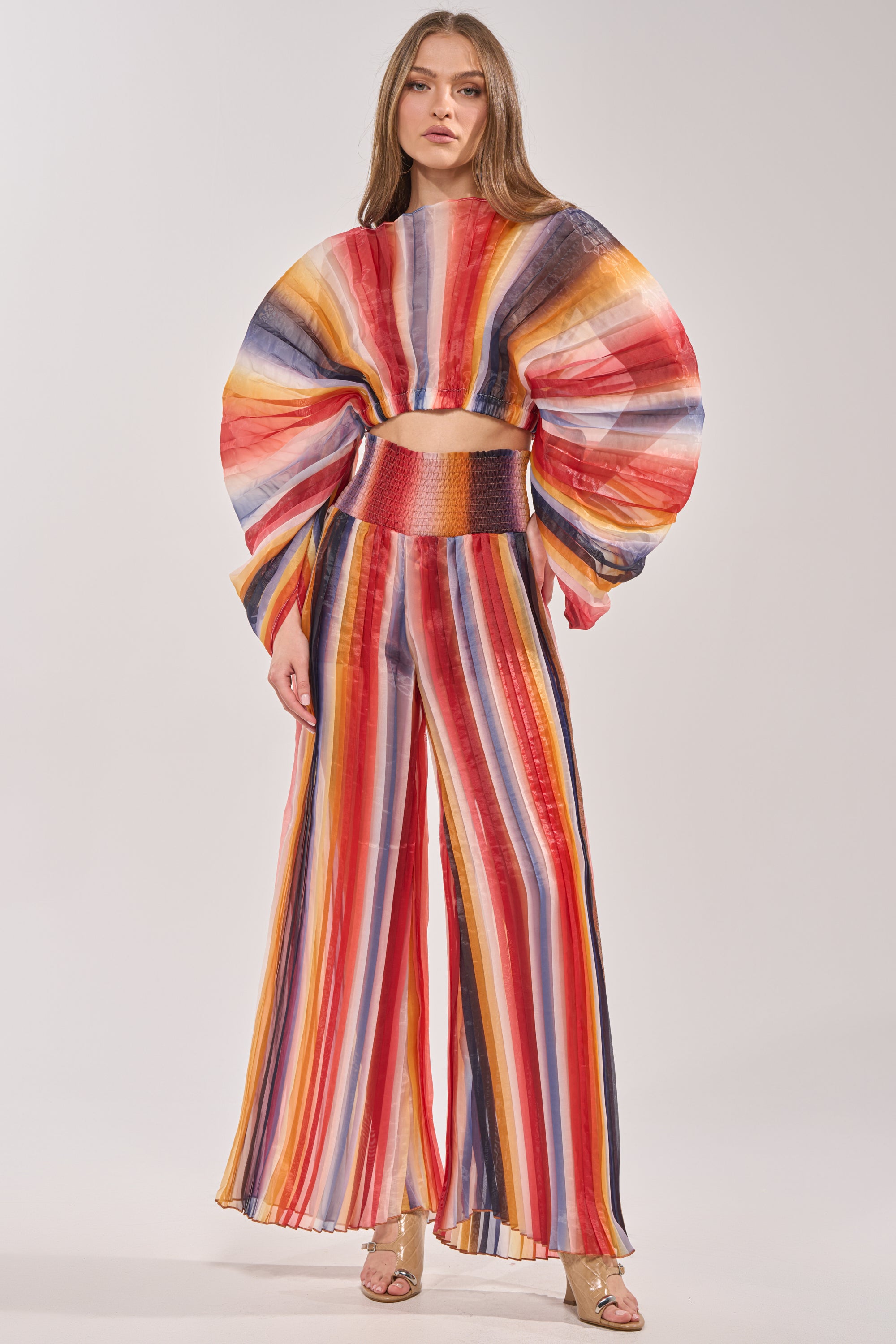 A woman in the FANNED OUT CHIFFON BLOUSE—long-sleeved, multicolor summer top—and vibrant striped wide-leg pants with gold high heels stands against a plain backdrop. 4