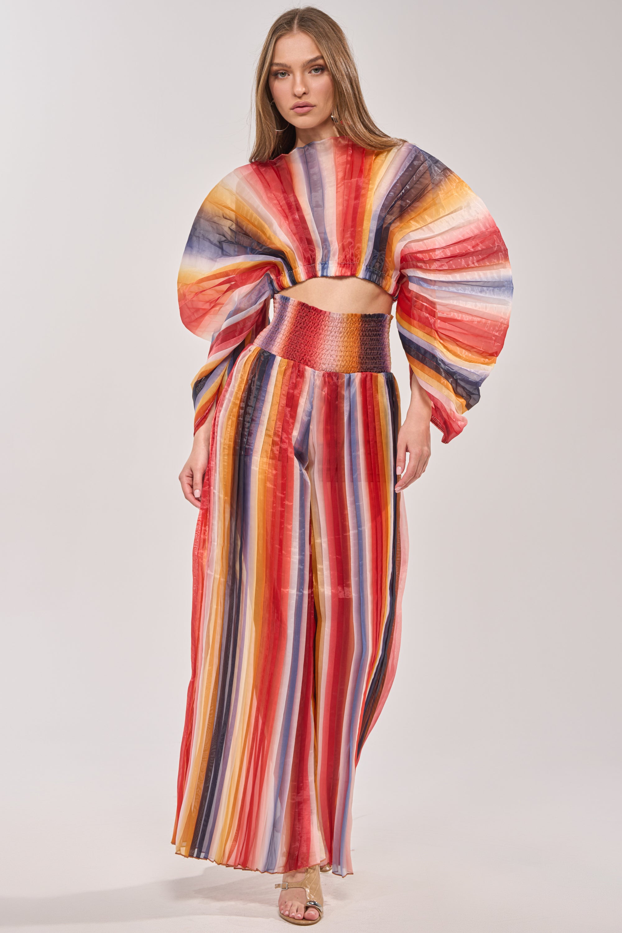 A woman stands against a plain background wearing the FANNED OUT CHIFFON BLOUSE—a vibrant, multicolor top with long wide sleeves and red, yellow, blue, and orange stripes—paired with a matching high-waisted skirt. 5