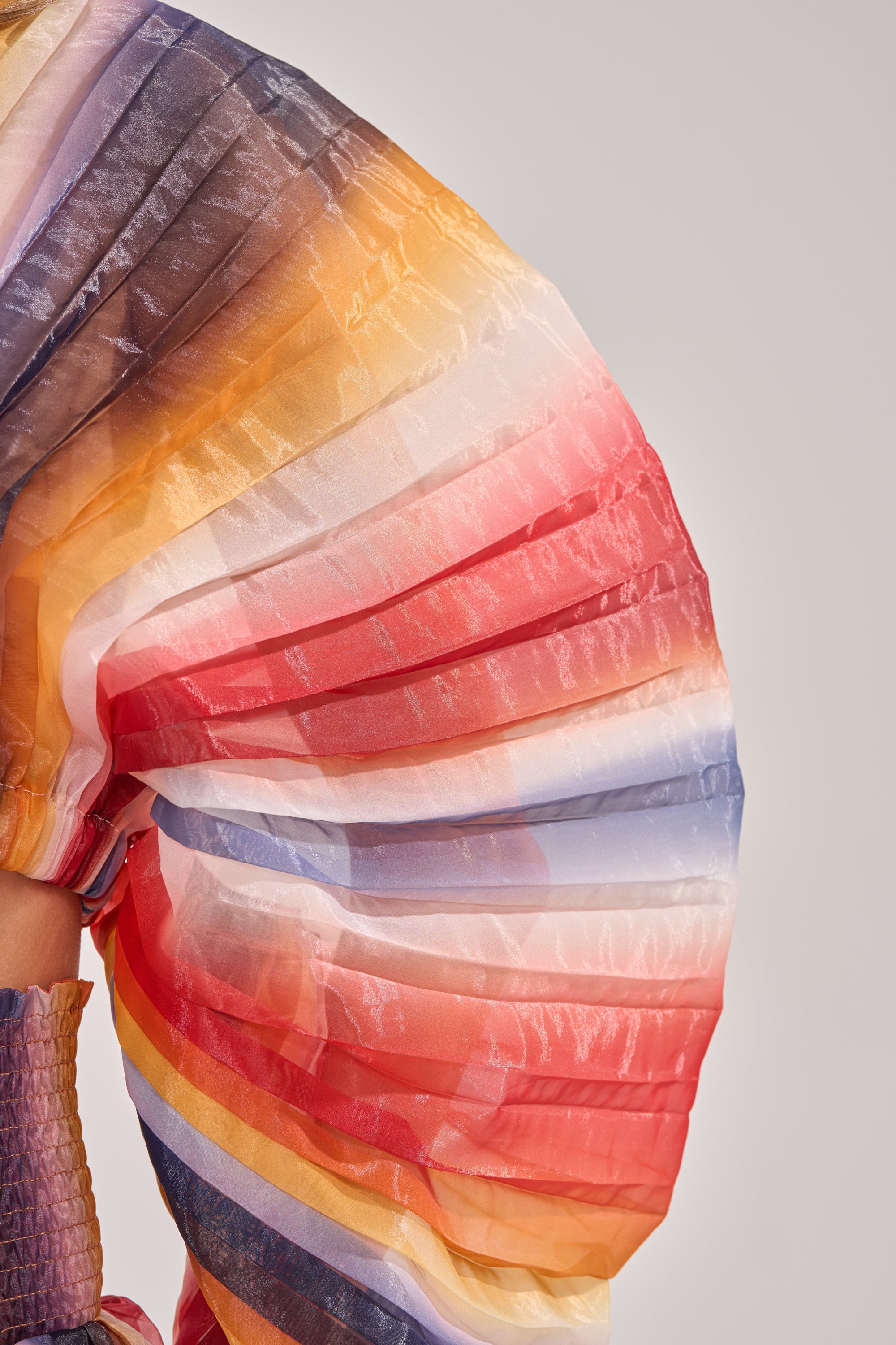 A close-up of the FANNED OUT CHIFFON BLOUSE highlights its vibrant, pleated fan sleeve with multicolor gradient hues of orange, red, yellow, blue, and white against a plain backdrop. 6