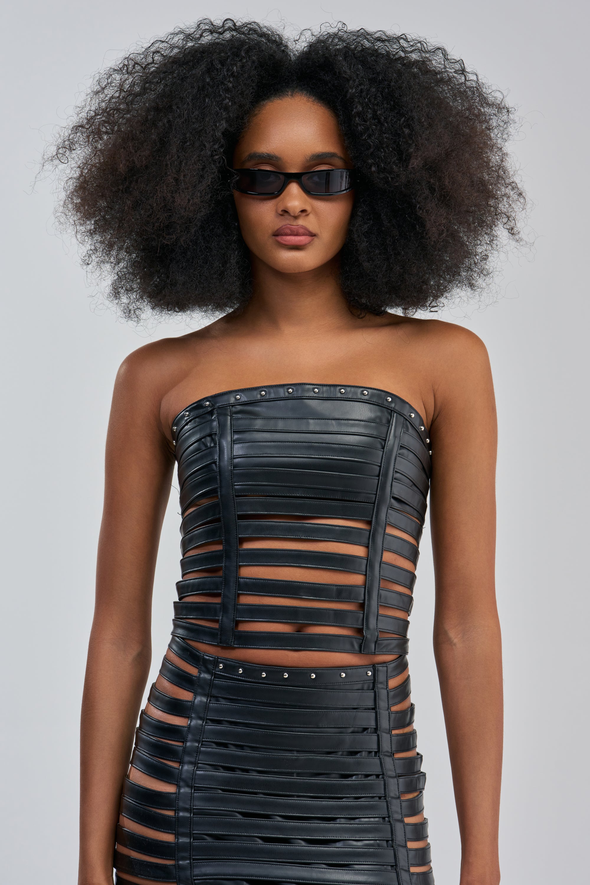 A woman with voluminous curly hair wears black sunglasses and the RAVE QUEEN PU TUBE TOP, paired with a skirt made of horizontal bands with cutouts, standing against a plain light background.
