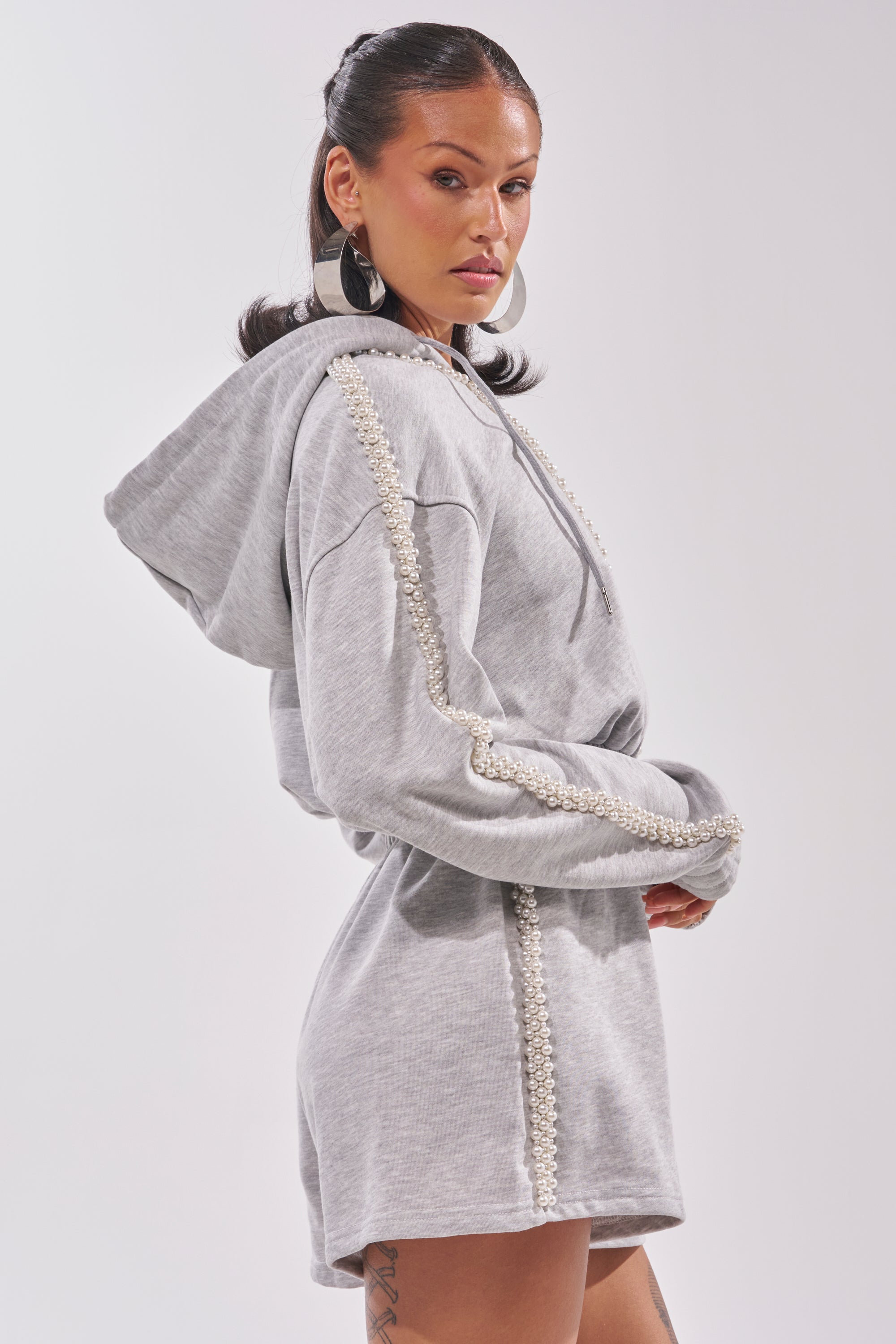 A woman poses sideways in the PEARLS FOR THE GIRLS FRENCH TERRY ROMPER, a hooded grey piece with pearl accents on the sleeves and sides. She styles it with hoop earrings, sleek hair, and shows off a tattoo on her upper thigh. 3