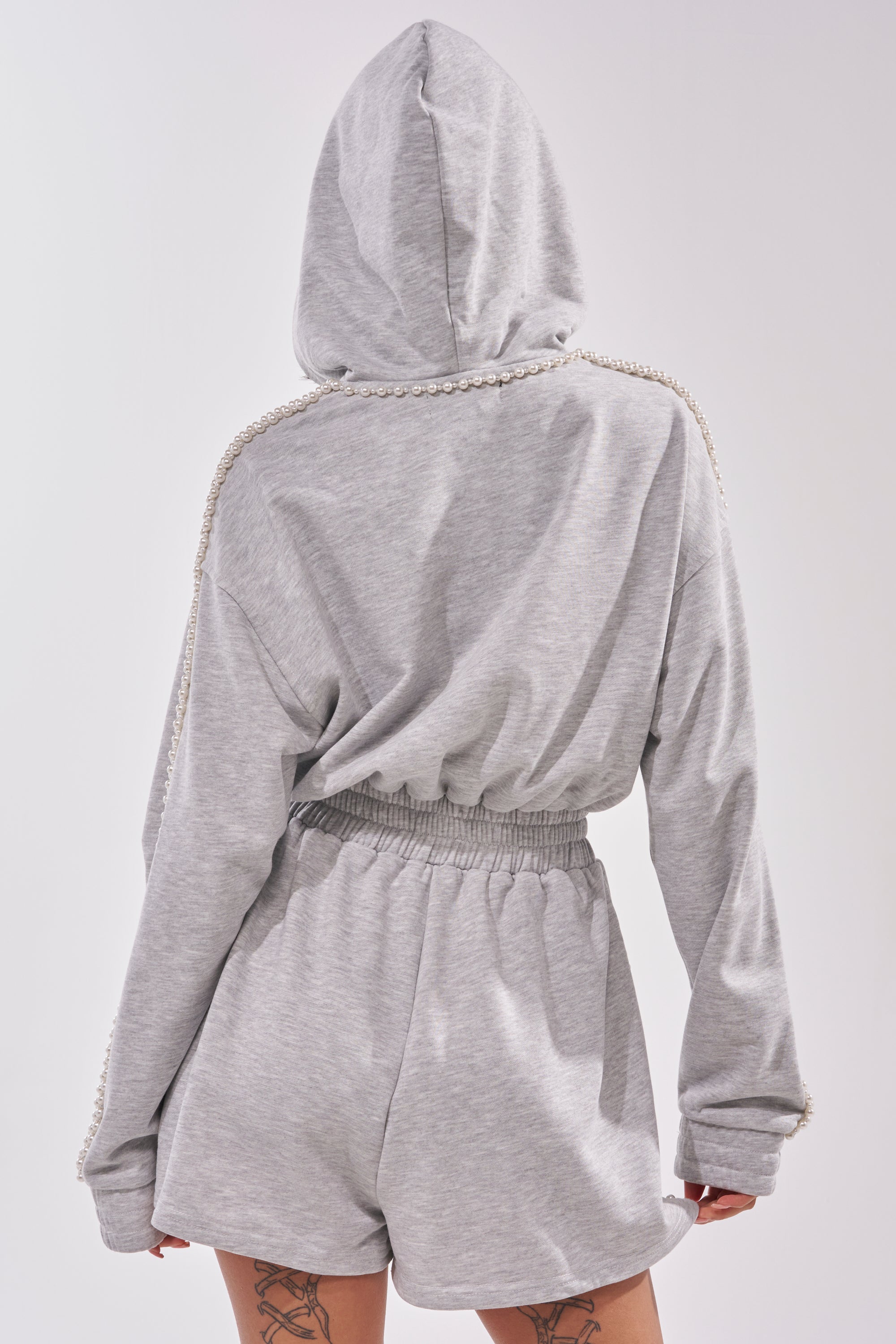 A person stands facing away, showing upper thigh tattoos while wearing the PEARLS FOR THE GIRLS FRENCH TERRY ROMPER—a grey hooded romper with pearl detailing on the shoulders. 4
