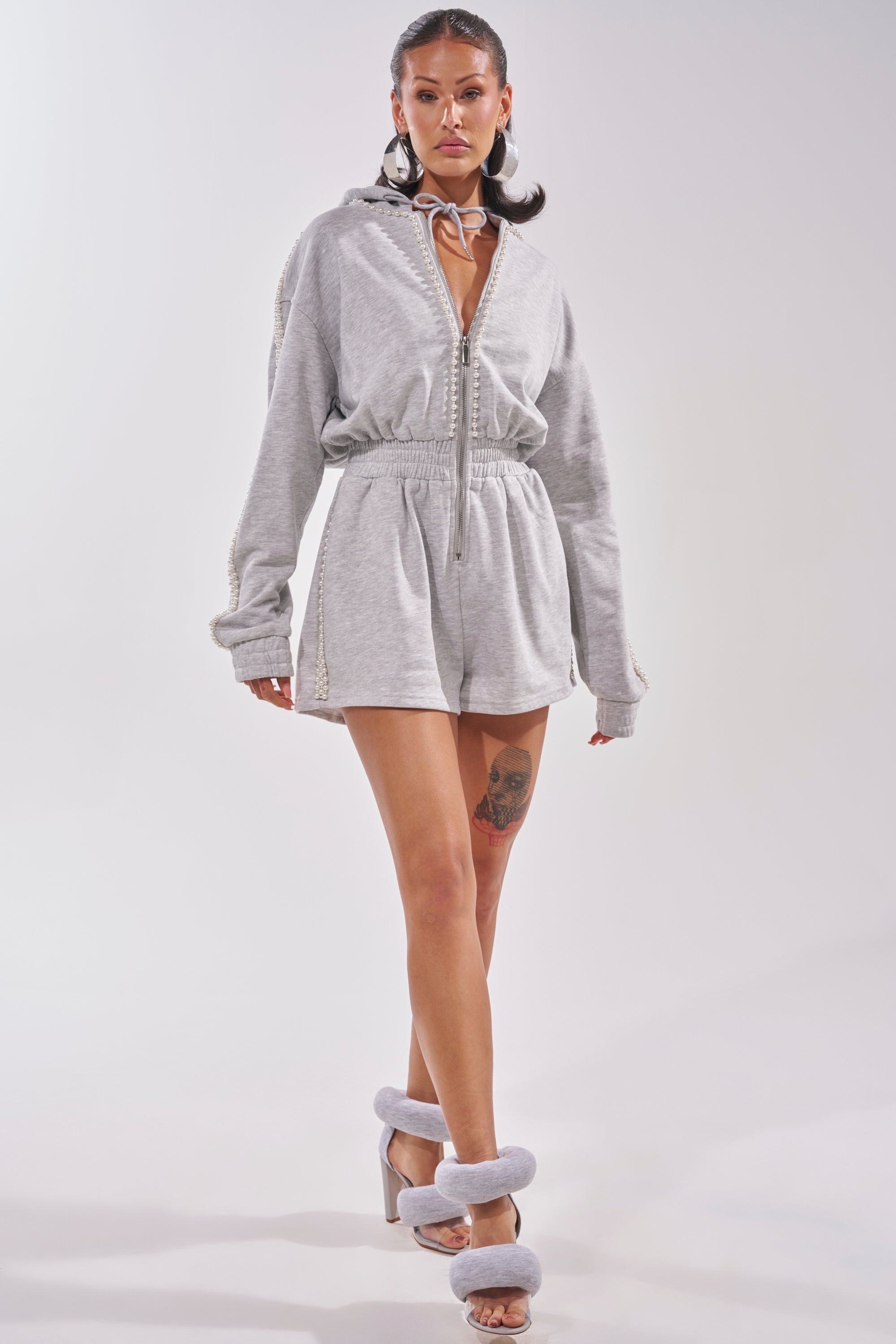 A woman stands confidently in the PEARLS FOR THE GIRLS FRENCH TERRY ROMPER—a light gray hooded, zip-up romper with a high waist and drawstring—paired with gray open-toe heels featuring fluffy straps, against a plain background. 6
