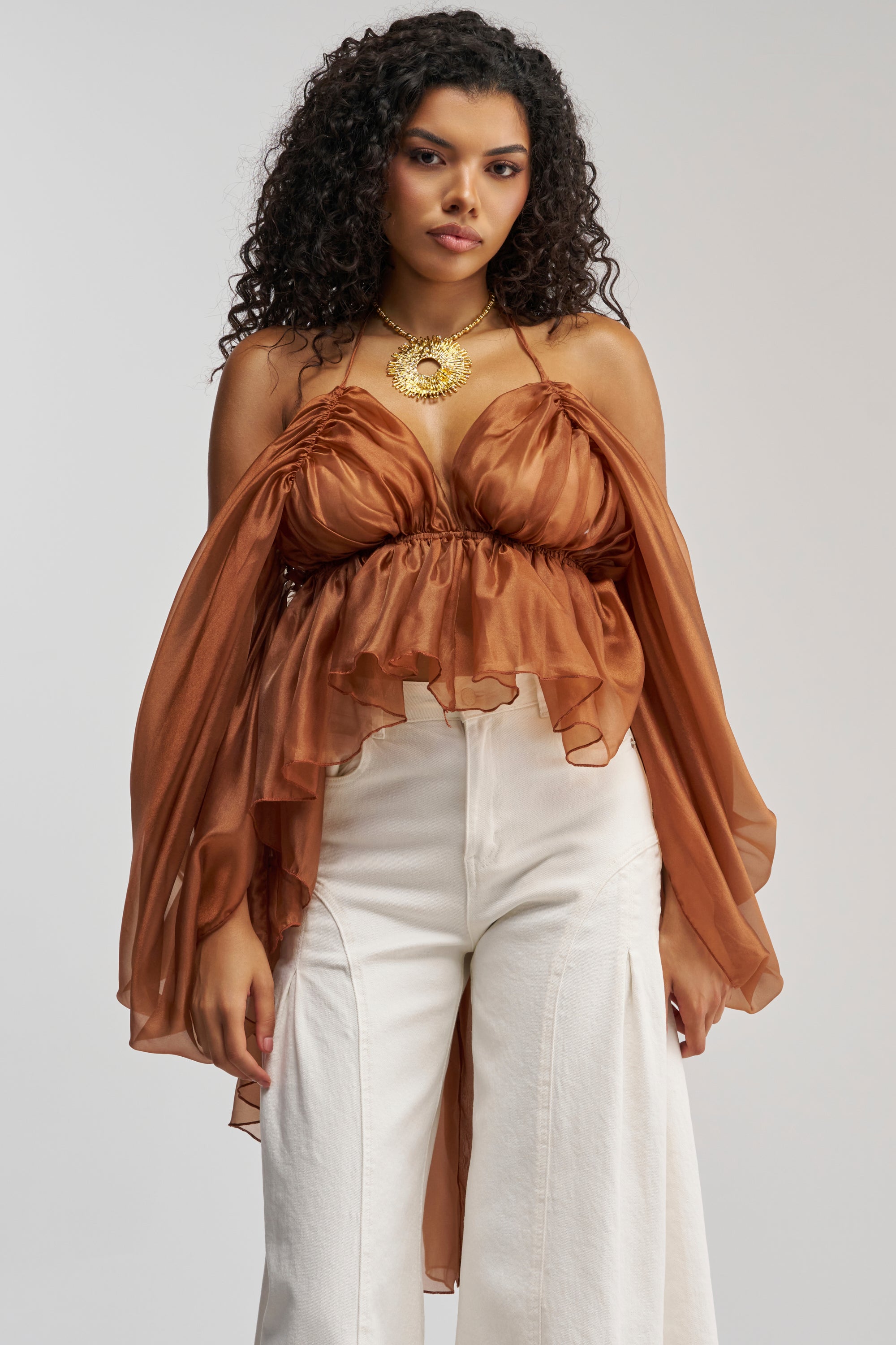 Styled against a light gray background, a woman with curly hair wears the FLY WITH ME CHIFFON BLOUSE—a rust-colored, open-back design featuring dramatic ruffled sleeves—paired with a statement necklace and high-waisted white pants, making it the perfect choice for standout occasion dressing.