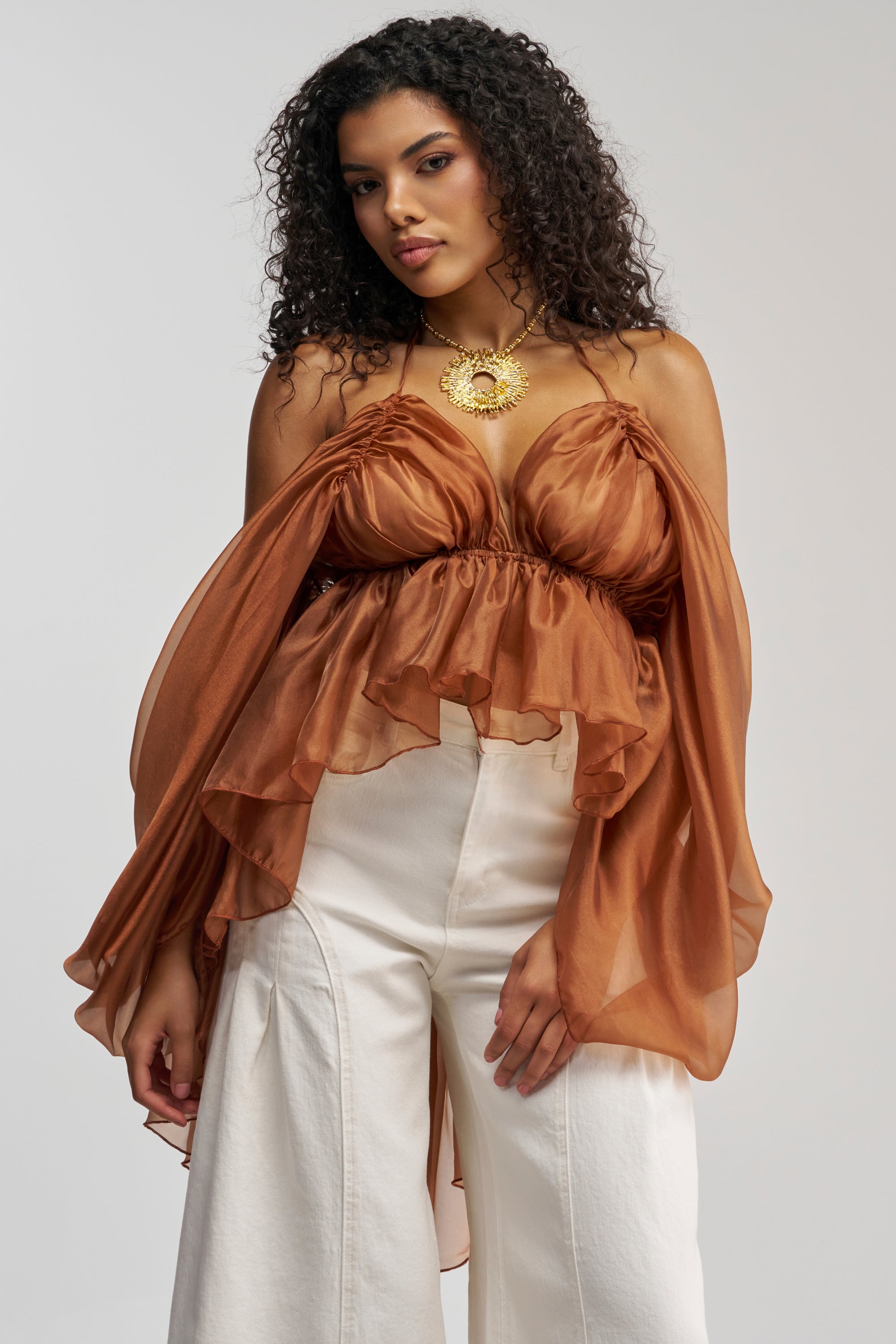 A woman with curly hair wears the FLY WITH ME CHIFFON BLOUSE—a brown sheer top with an open-back design and dramatic, flowing sleeves—styled with white wide-leg pants and a bold gold sun-shaped necklace, creating an elegant statement look against a plain background.
