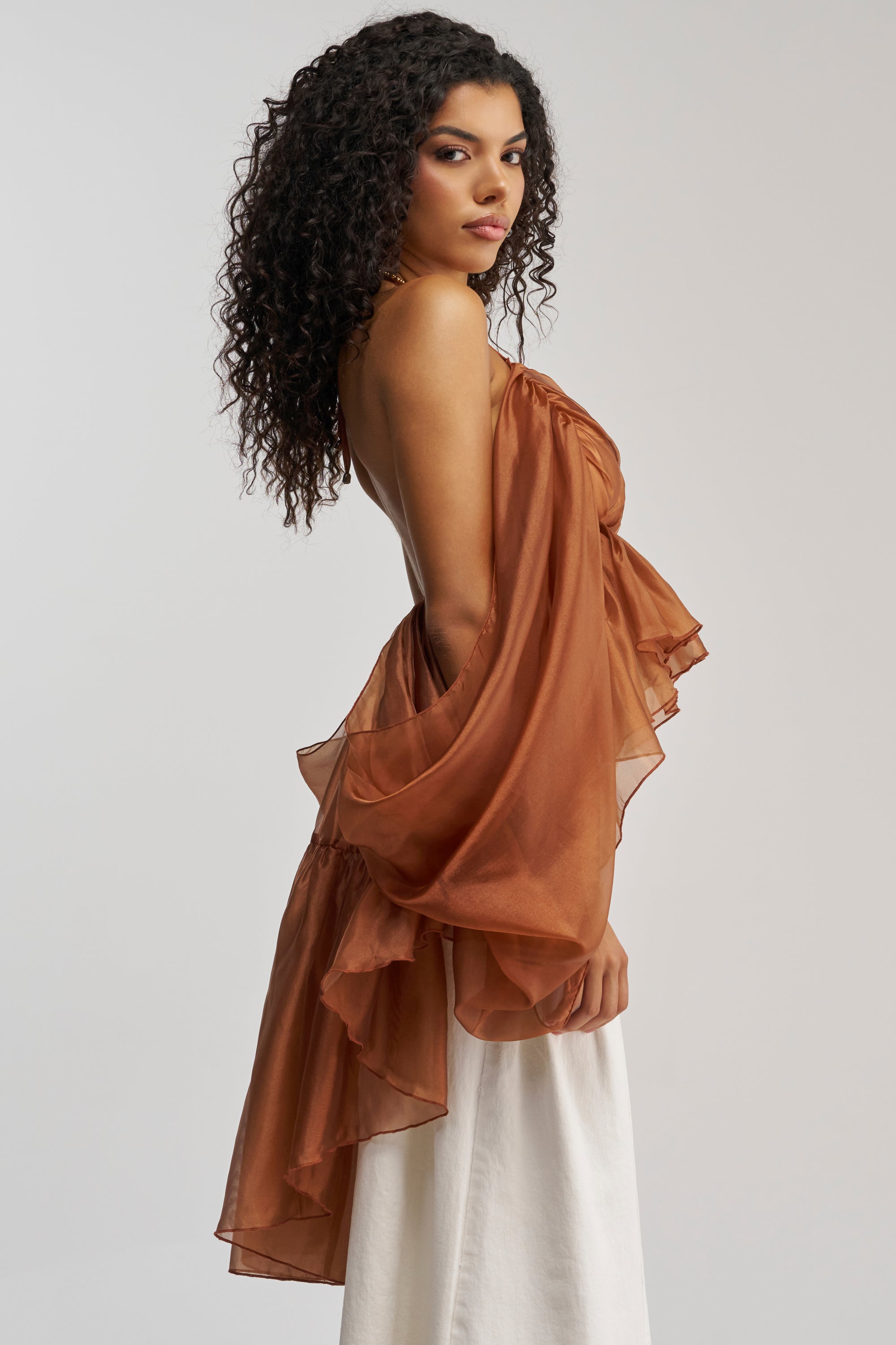 A woman with curly hair showcases the FLY WITH ME CHIFFON BLOUSE—a brown, sheer piece featuring ruffled, off-the-shoulder details—paired with a long white skirt for an elegant statement occasion look as she poses sideways against a plain light background.