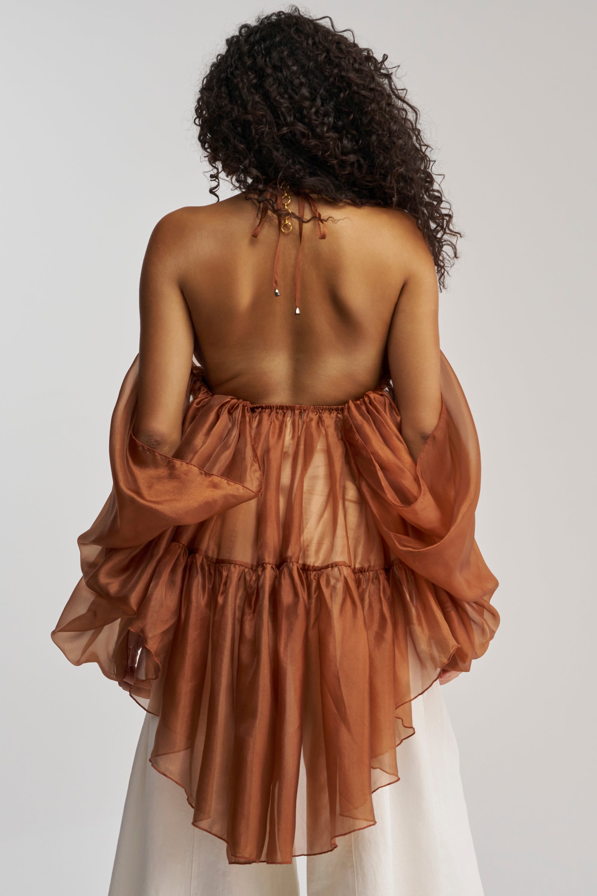 A person with curly hair is seen from the back, wearing the FLY WITH ME CHIFFON BLOUSE—a brown sheer blouse featuring an open-back design with puffy sleeves and a tiered silhouette—paired with white pants, perfect for making a statement at any special occasion.