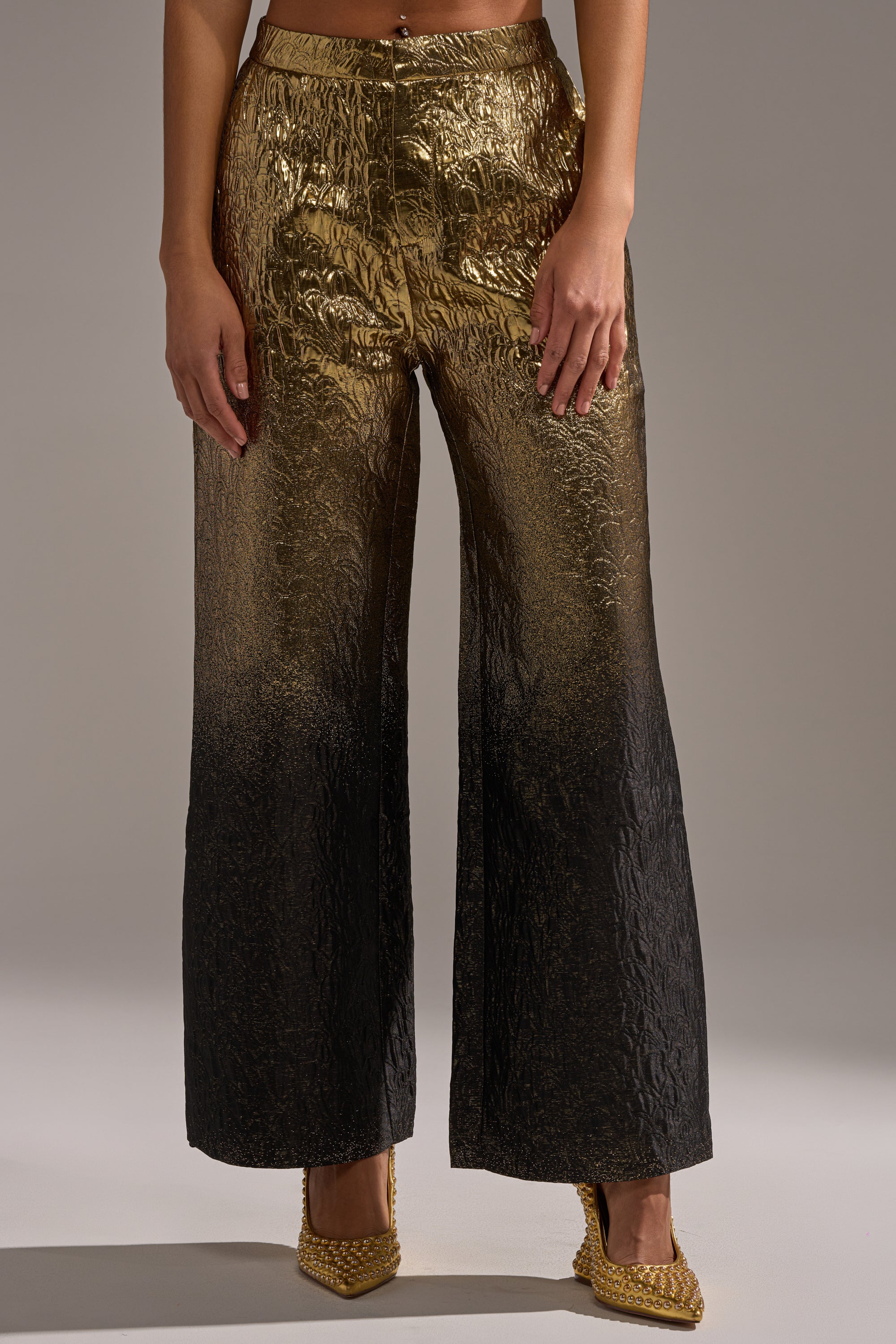 A person wears the ALL ABOUT THE DRAMA TROUSER—high rise, festive pants with a metallic gold-to-black ombre effect and textured fabric—paired with gold pointed-toe heels. The upper body is not visible.