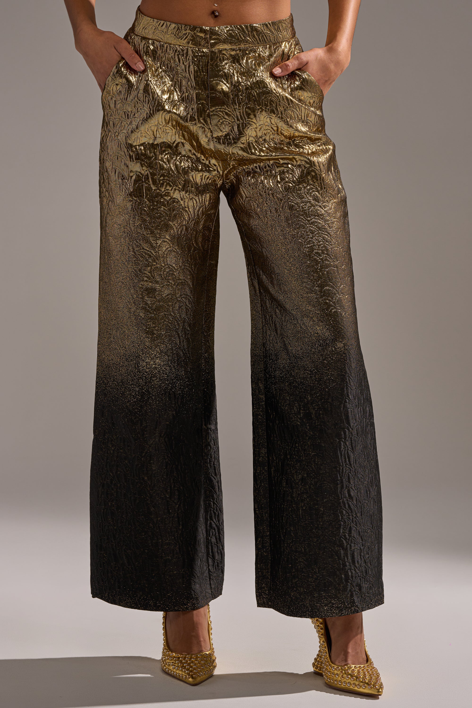 Wearing the ALL ABOUT THE DRAMA TROUSER, featuring a high-rise fit, gold-to-black ombré pattern, and textured finish. Hands are in pockets and paired with pointed gold heels; upper body is cropped out.