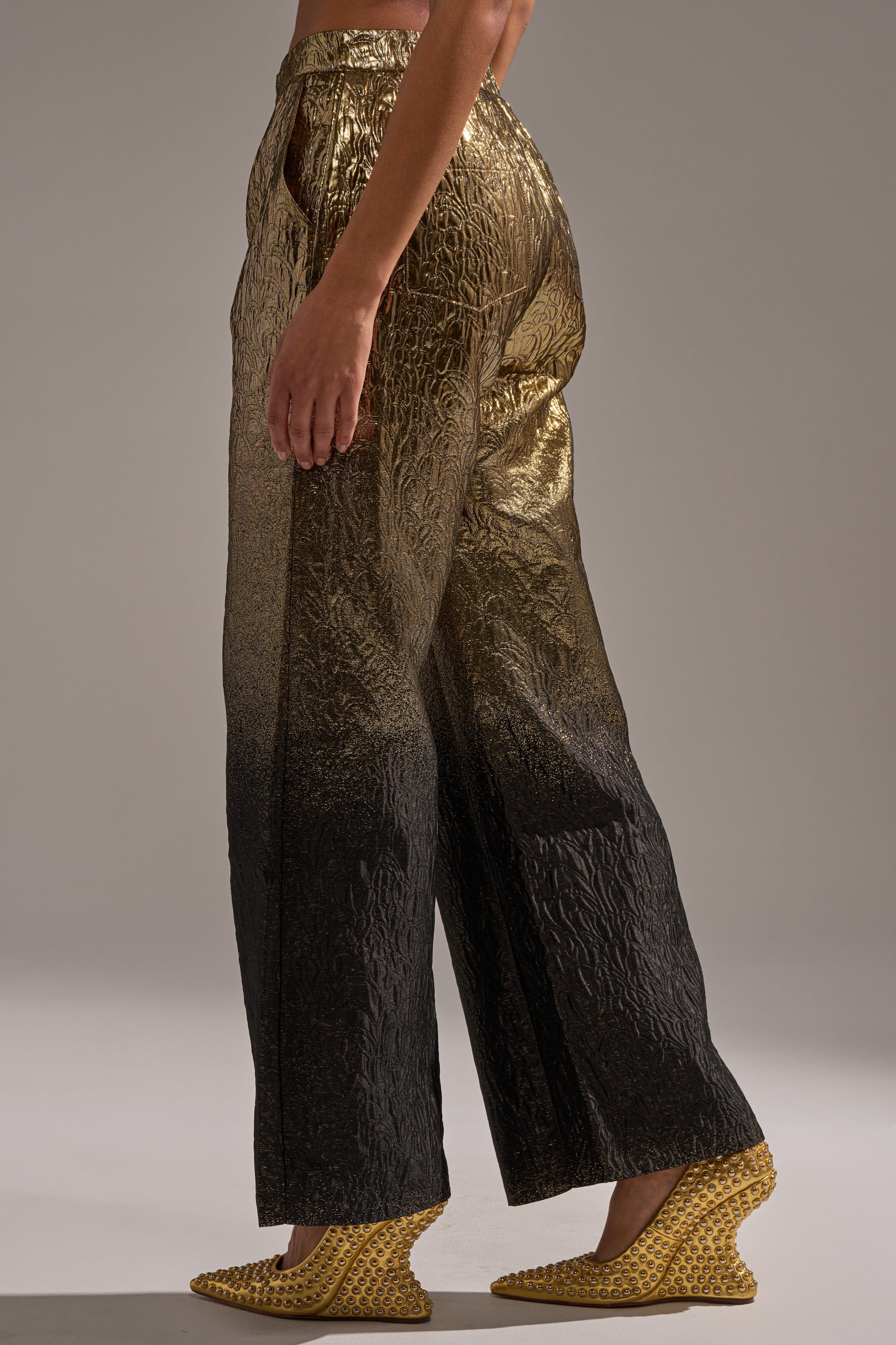 A model showcases the ALL ABOUT THE DRAMA TROUSER—high-rise, wide-leg pants with a metallic gold to black ombré textured finish—styled with yellow wedges against a plain background.