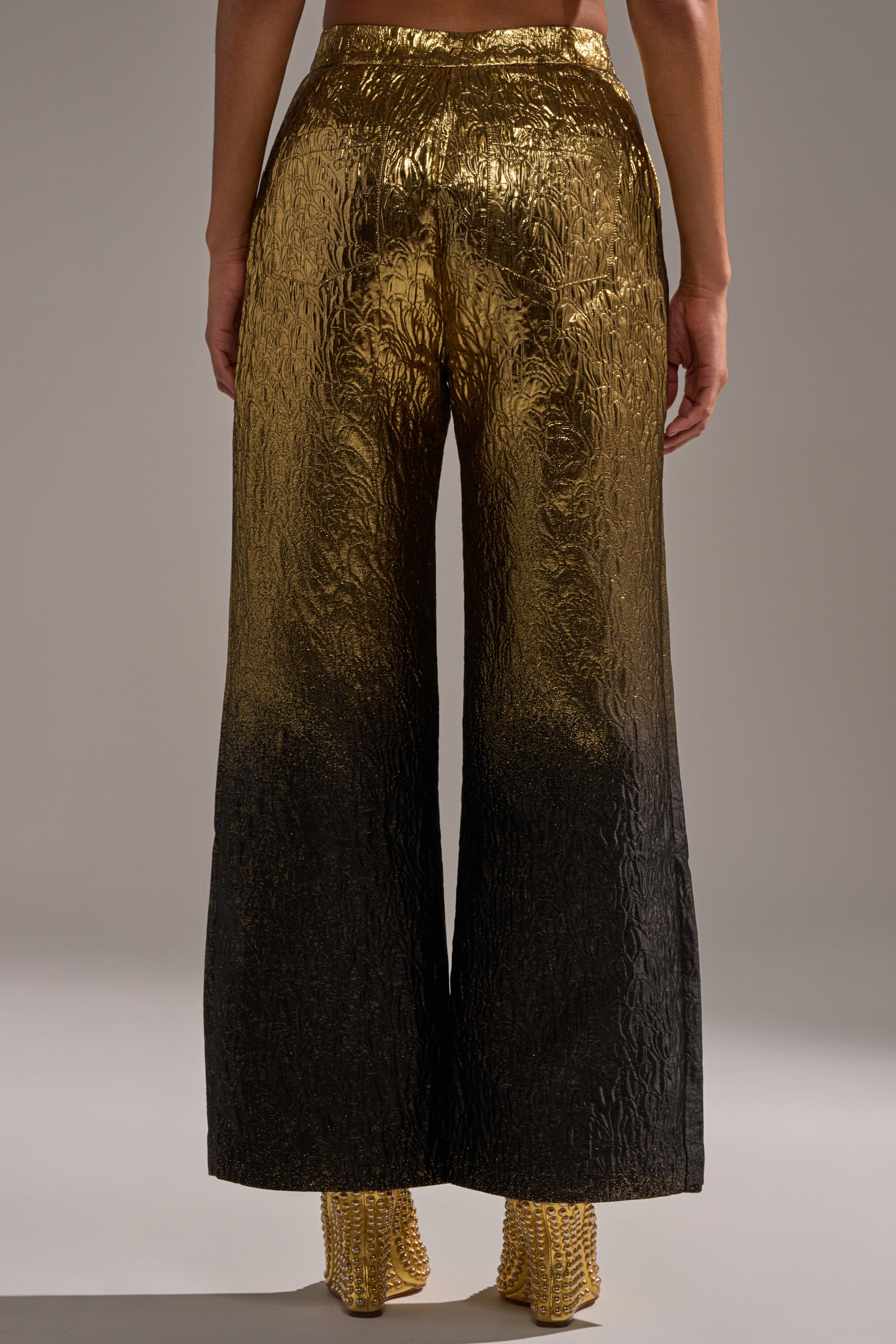 A person stands facing away, wearing the ALL ABOUT THE DRAMA TROUSER—high-rise, wide-leg pants with a textured metallic gold finish fading to black—paired with gold, textured heeled shoes.