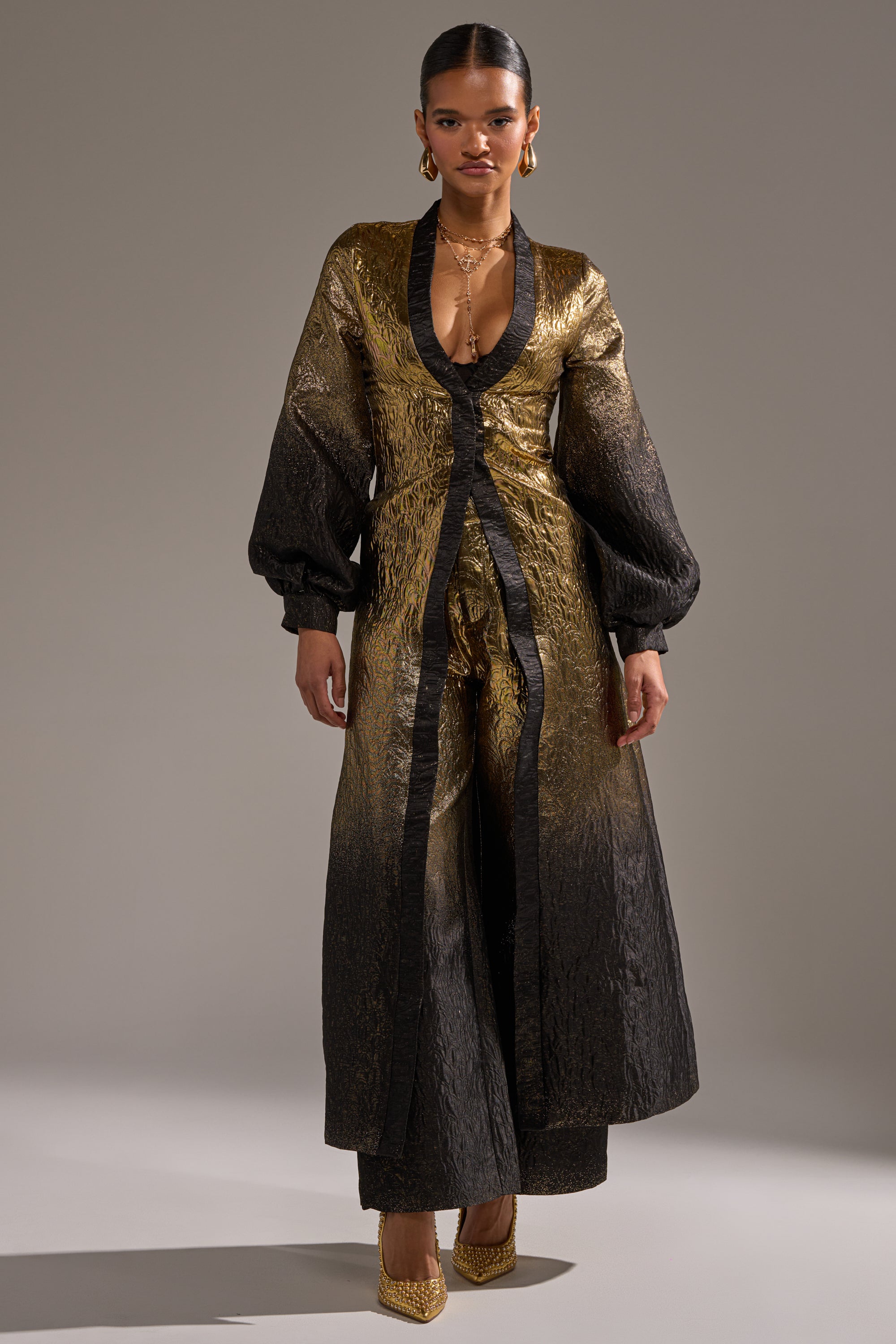A woman poses in high-rise, wide-leg ALL ABOUT THE DRAMA TROUSER pants with gold heels, gold earrings, layered necklaces, and a textured gold and black gown featuring a deep V-neck and dramatic long sleeves against a gray background.