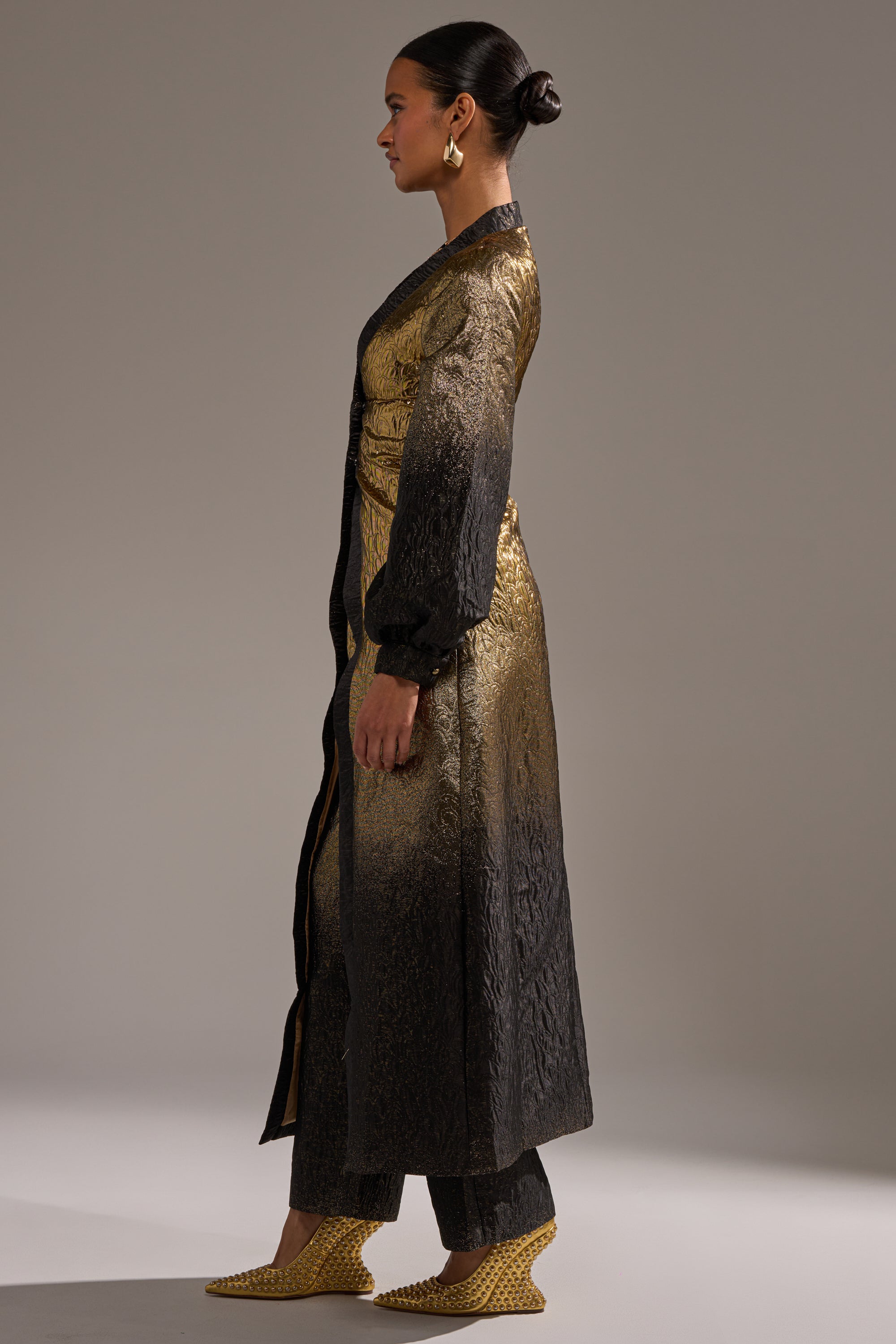 A woman in profile wears the ALL ABOUT THE DRAMA OVERSIZED BLOUSE in gold-to-black ombre, with matching pants, gold shoes, hair in a neat bun, and gold hoop earrings against a plain background.