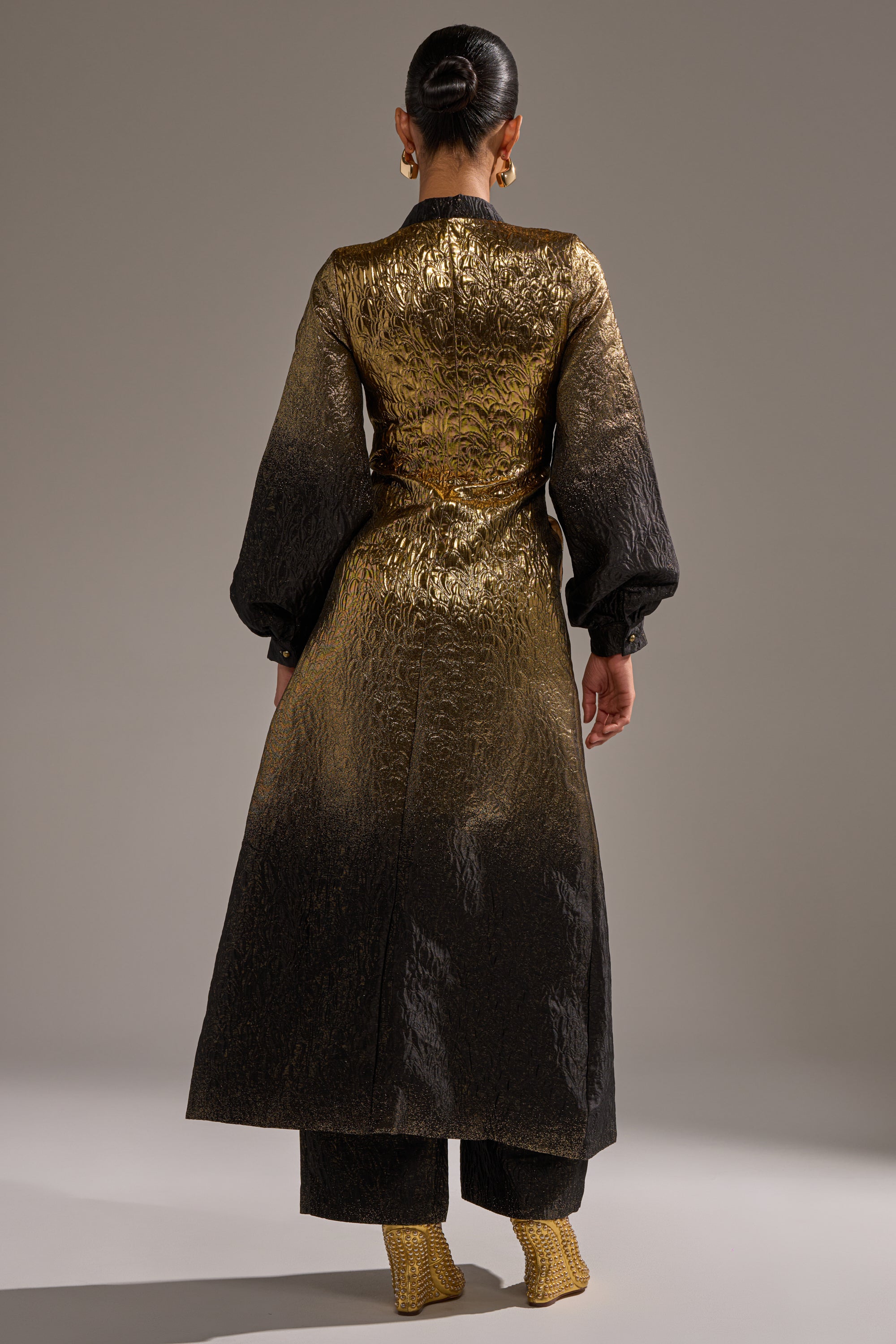 A person stands facing away, wearing the ALL ABOUT THE DRAMA OVERSIZED BLOUSE—a shimmering, metallic longline top with wide sleeves—paired with matching textured pants, heeled shoes, gold hoop earrings, and their dark hair in a sleek bun.