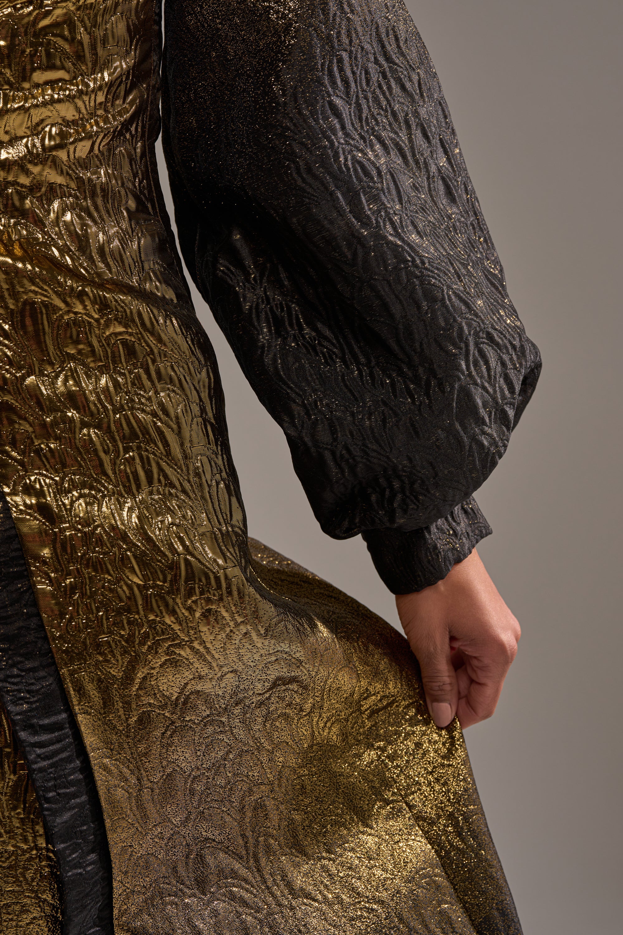A person models the ALL ABOUT THE DRAMA OVERSIZED BLOUSE, featuring a metallic gold and black pattern with voluminous sleeves, holding the fabric at their side against a plain gray background.