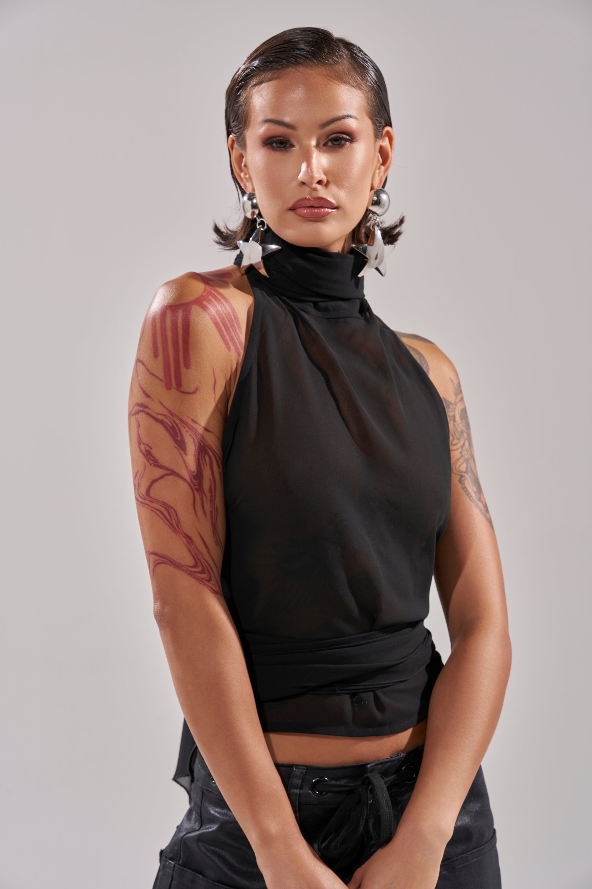 A person with slicked-back hair models the GOT IT FROM YOU BLOUSE, paired with star-shaped earrings and dark pants. Red and black tattoos are visible on their arms as they pose confidently against a plain light background. 0