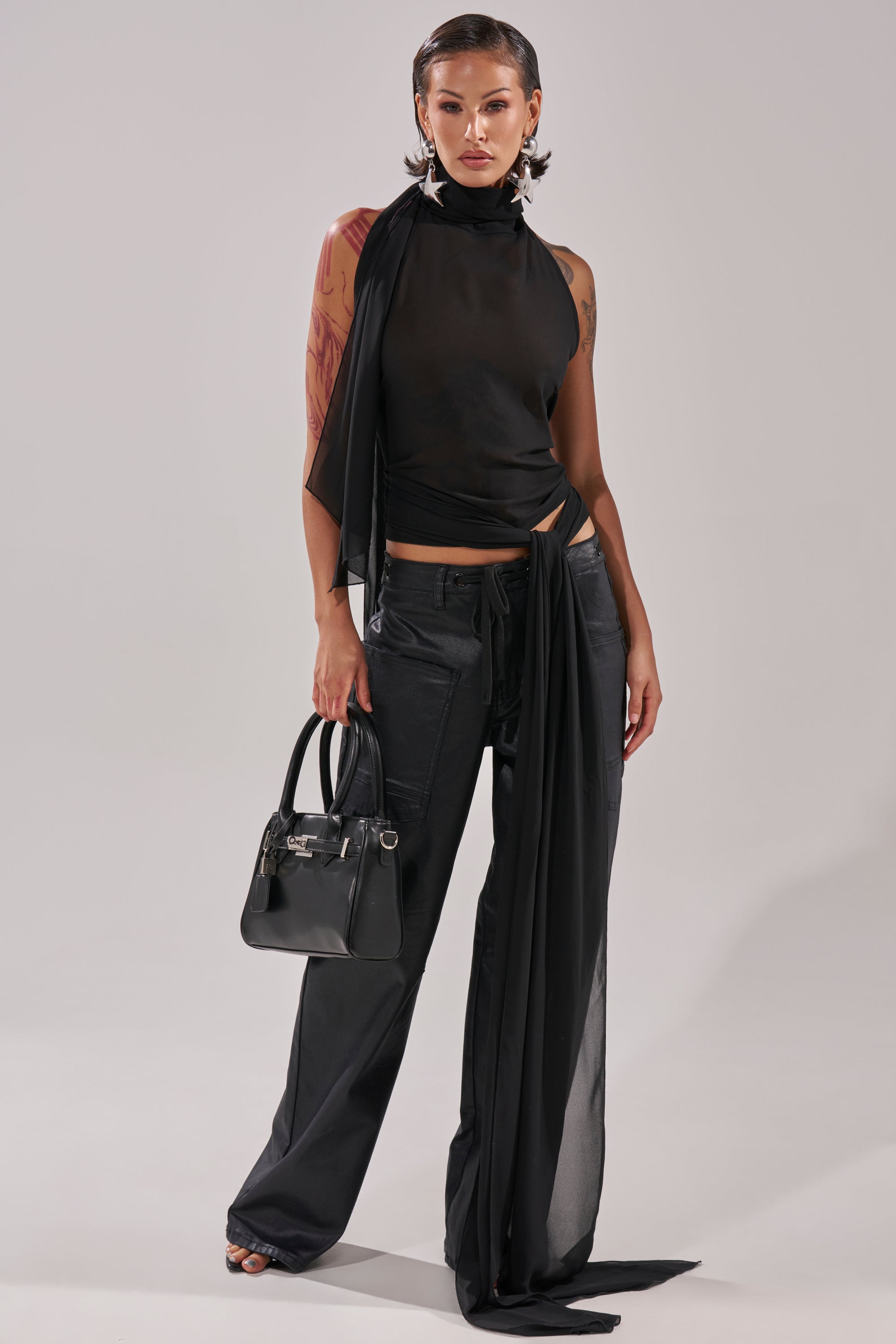 A woman stands confidently in black wide-leg pants and the GOT IT FROM YOU BLOUSE—a black halter wrap top with draped fabric—paired with large star earrings, a stylish black handbag, and wet-look hair against a plain light background. 2