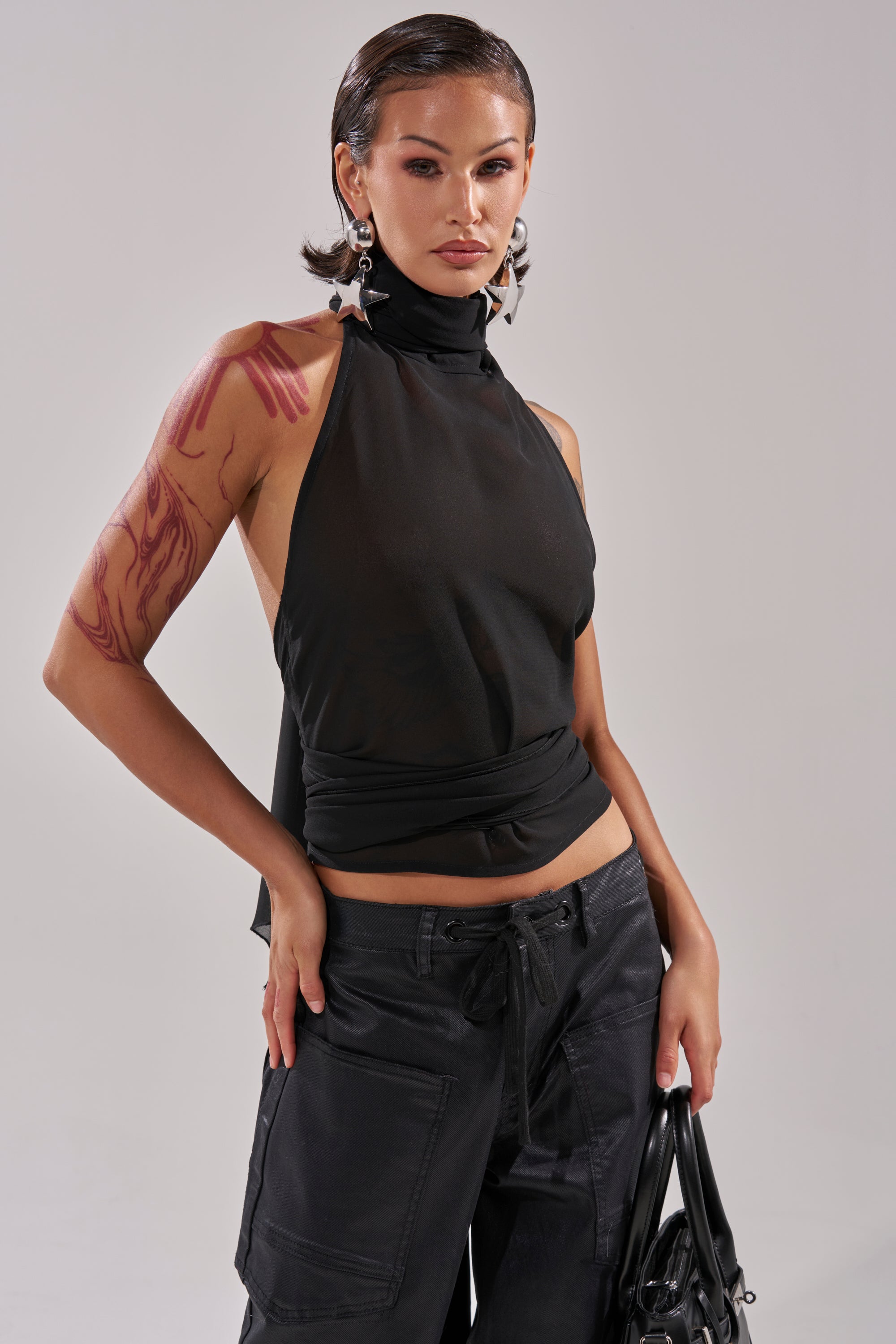 A woman with slicked-back hair and star earrings wears the GOT IT FROM YOU BLOUSE in black, paired with black pants. She displays a large red tattoo on her left arm, holds a black bag, and poses confidently against a plain background. 4
